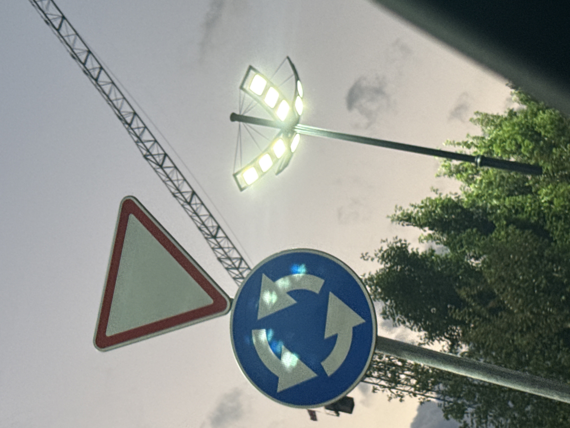 The image shows a street scene with traffic signs and a lamppost against a cloudy sky. In the foreground, there is a blue circular sign with white arrows indicating a roundabout. Above it, a triangular yield sign with a red border is visible. To the right, a modern lamppost with multiple lights illuminates the scene. In the background, a construction crane is partially visible, and trees can be seen below. The overall lighting suggests it is dusk or overcast. The context of Tbilisi, Georgia, is not directly evident from the visual elements of the image itself.