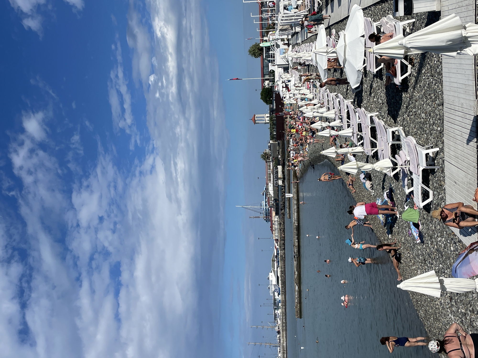 The image depicts a bustling beach scene in Sochi, Russia, likely during the summer season. The foreground is dominated by a pebble beach covered with numerous white lounge chairs and umbrellas. Several people are scattered across the beach, with some sunbathing on the loungers, while others are standing or walking.
In the middle ground, the calm sea is populated by many swimmers, including adults and children, enjoying the water. Some individuals are wading near the shore, while others are further out. The waterline is clearly defined, with the dark pebbles transitioning to the clear water.
The background reveals the Sochi seascape and cityscape. A harbor with various boats and ships is visible, suggesting the area's maritime importance. Buildings with distinct architectural styles line the waterfront, including what appears to be a tower with a red-tiled roof. Tall masts of sailing vessels and industrial structures are also present, contributing to the dynamic scenery. The sky is a mix of blue with scattered white clouds, indicating fair weather conditions. The overall impression is one of a popular tourist destination, with a vibrant atmosphere of recreation and leisure.