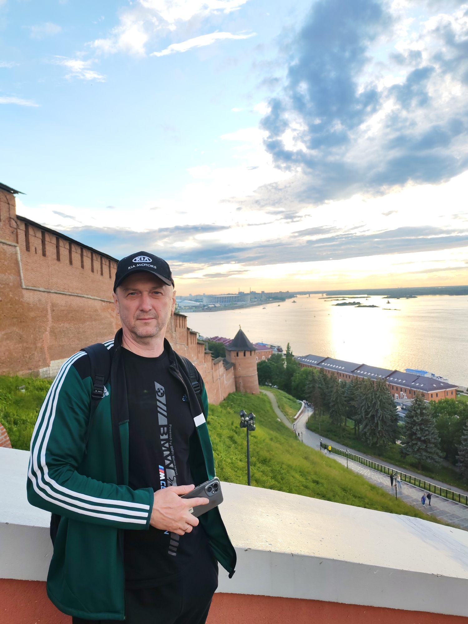 A man wearing a black baseball cap with "KIA MOTORS" text and a green and white Adidas jacket stands on a wall overlooking a river and a city at sunset. He is also wearing a black t-shirt with "BMW MOTORSPORT" text. He holds a smartphone in his right hand. The background features a large river, a bridge, and buildings. The sky is filled with dramatic clouds, and the sun reflects on the water, indicating it is late afternoon or early evening. The man appears to be a tourist, posing for a photo while enjoying the scenic view of Nizhny Novgorod, Russia. The ancient brick fortress walls, visible to the left, suggest the location is likely the Kremlin.