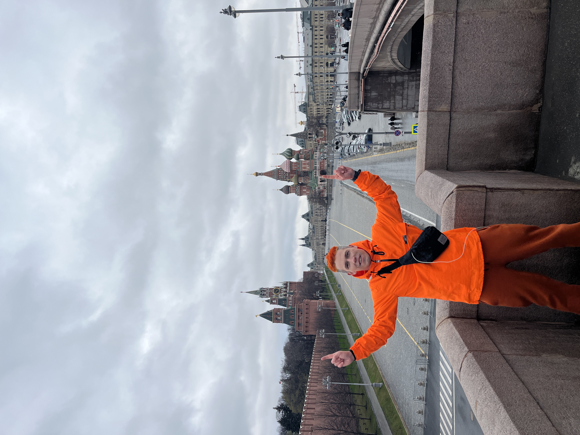 A man wearing an orange jacket and pants stands on a stone bridge in Moscow, Russia, gesturing with both hands. He has red hair and a black crossbody bag. Behind him is a wide view of Red Square, featuring the Kremlin walls, St. Basil's Cathedral, and other buildings in the distance. The sky is overcast and gray. The street below the bridge appears wet.