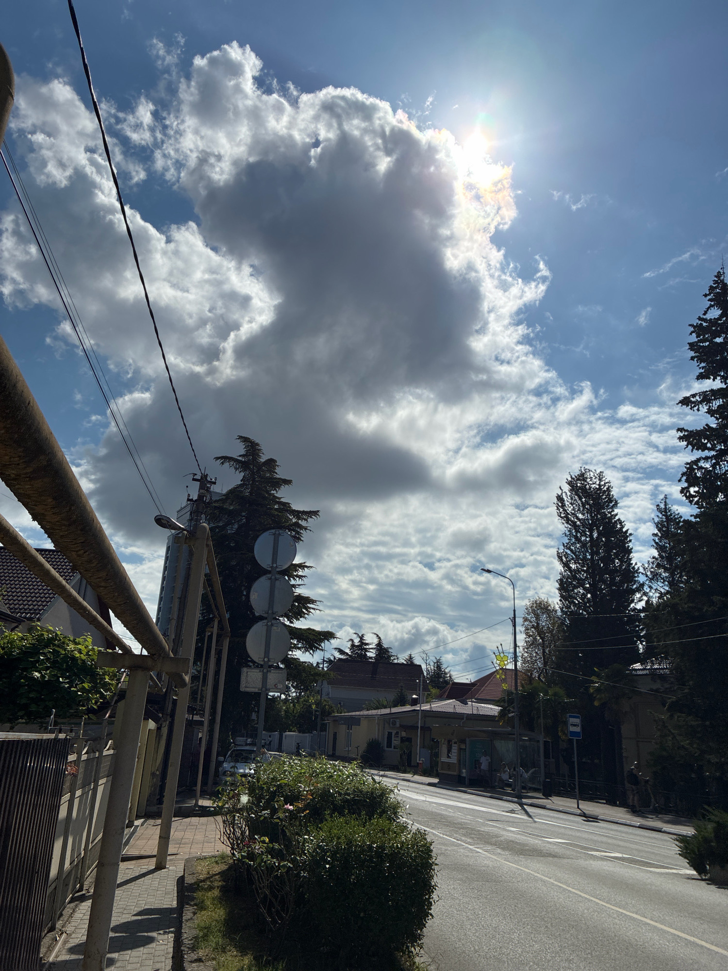 The image shows a street scene in Sochi, Russia, under a partly cloudy sky with the sun shining brightly. The foreground features a paved sidewalk bordered by a metal fence and lush green bushes. A large, metal pipe runs diagonally across the upper left of the frame. Beyond the sidewalk, a road curves to the right, with a bus stop on the opposite side. Several people are visible at the bus stop, some sitting and others standing. Tall evergreen trees line the right side of the road and frame the background. In the distance, buildings with red-tiled roofs are visible. Power lines stretch across the sky, and a street lamp stands near the bus stop. The overall impression is of a typical urban environment on a sunny day.
