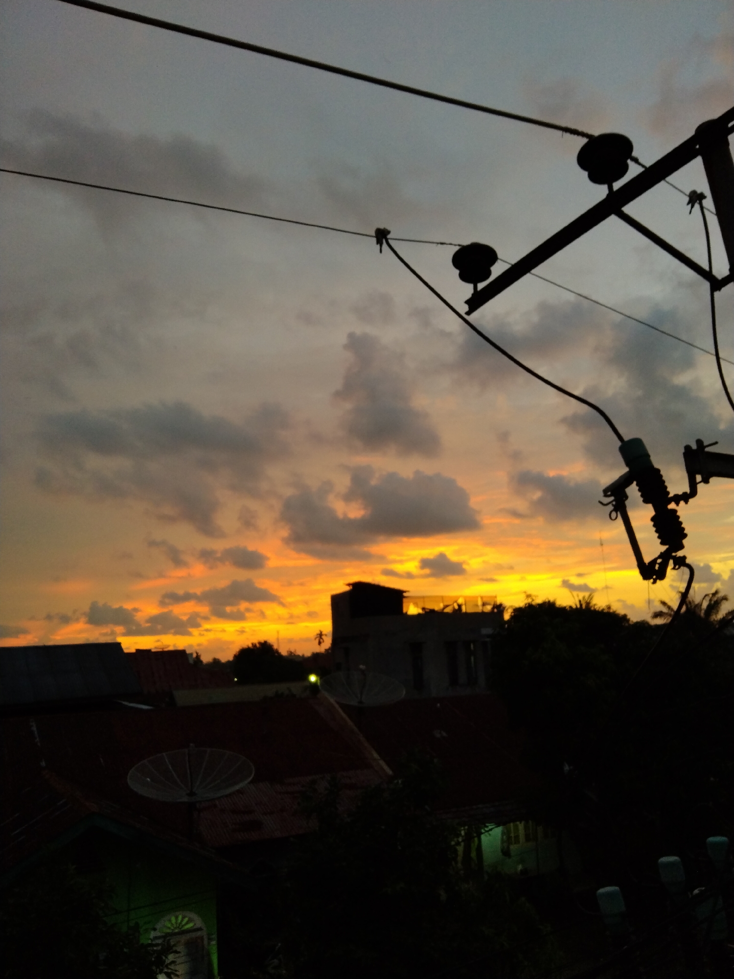 The image captures a vibrant sunset over a residential area in Bireuenmeunasahdayah, Indonesia. The sky dominates the upper and mid-sections, displaying a dramatic gradient of colors. At the top, the sky is a muted gray-blue, transitioning to darker, heavy clouds in the mid-sky. Below these clouds, the horizon glows with intense shades of orange and yellow, indicating the sun is setting.
Prominently featured in the upper right is the silhouette of a utility pole with multiple wires extending across the frame. The pole's insulators and hardware are distinctly visible against the colorful sky, as are several power lines running horizontally across the entire image.
Below the horizon, the dark silhouettes of numerous houses and buildings are visible. One roof in the lower left features a large, dark satellite dish. Trees and foliage are scattered among the buildings, appearing as dark masses against the bright sky, with hints of tropical palm trees. A faint greenish light emanates from a window or porch of a house in the lower left quadrant.
The scene depicts the tranquil late afternoon or early evening as daylight fades. No people are visible, and there are no specific activities taking place other than the natural progression of the day. There is no visible text in the image. The cloudy weather adds texture to the colorful sunset.