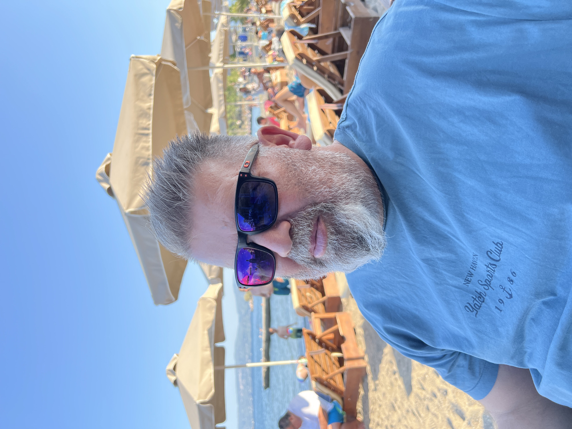 A medium shot captures a man in sunglasses and a blue t-shirt at a beach in Eretria, Greece. He is seated on the sand, with beach umbrellas and lounge chairs visible in the background, along with the sea and other beachgoers. The man's t-shirt features a graphic with the text "NEW HAVEN Yatch Sports Club 19 ¢ 86". The sky is clear and blue.