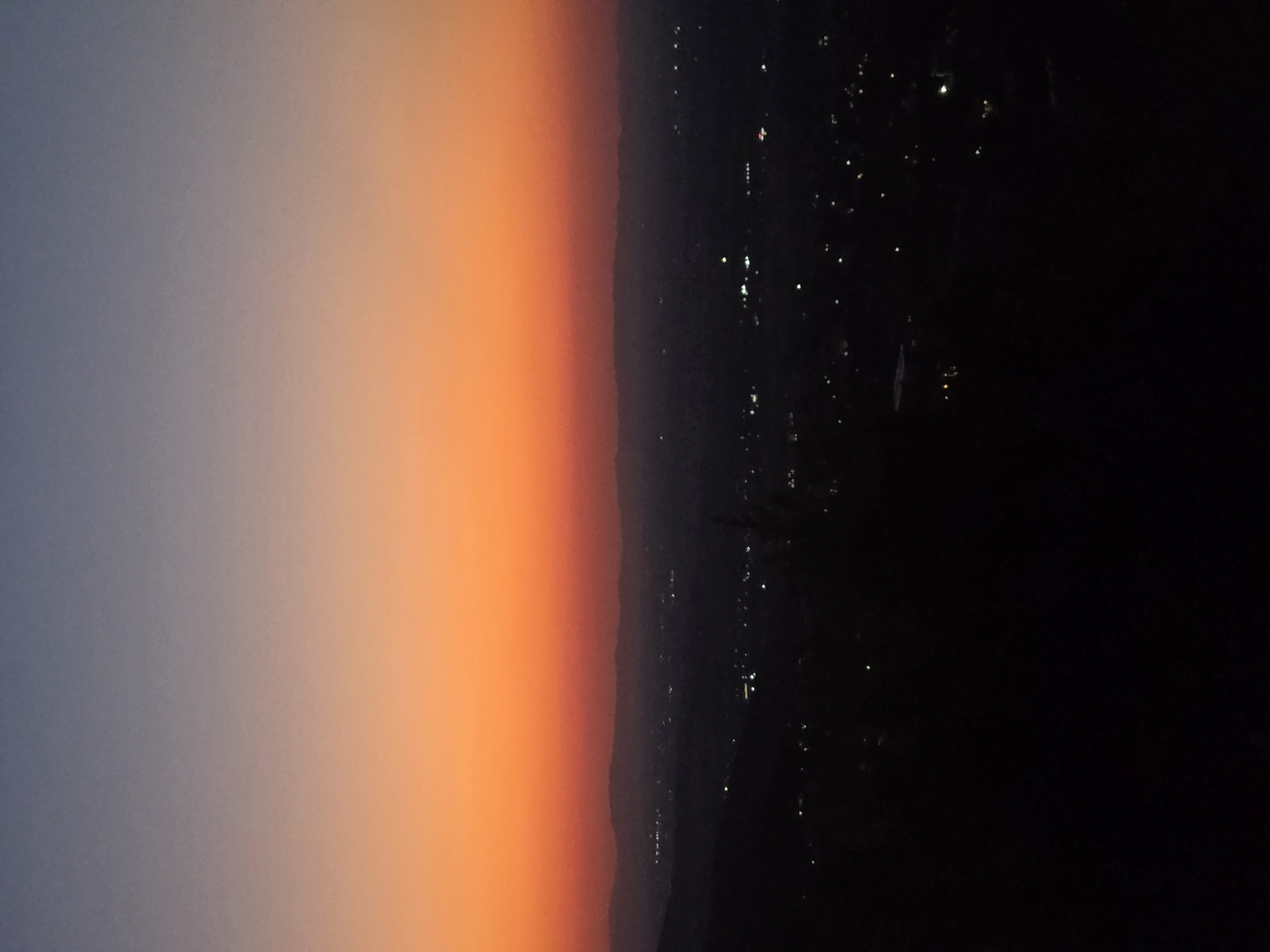The image depicts a panoramic view of Markovo, Bulgaria, during twilight. The sky transitions from a deep blue at the top to a vibrant orange hue closer to the horizon. Below the sky, silhouetted hills form a dark line against the gradient. The landscape is dotted with numerous small, glowing lights, indicative of a town or city at night. The foreground is entirely black, obscuring any details of the immediate surroundings.