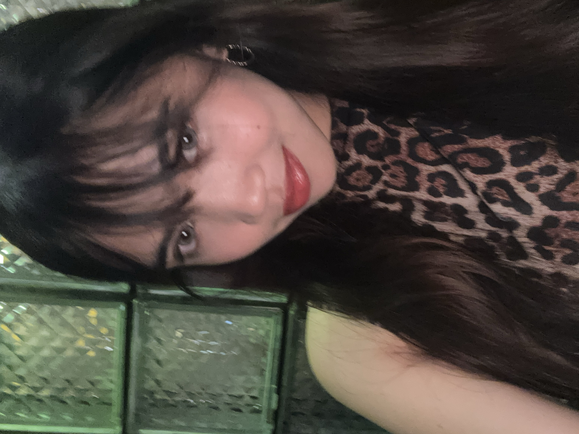 A young woman with dark hair and bangs is captured in a close-up selfie. She is wearing a leopard-print halter top and a hoop earring in her right ear. Her lips are painted red, and she has a mole on her left cheek. The background is a textured green glass block wall, common in many establishments in Davao City, Philippines, suggesting the photo was taken indoors in a bar, restaurant, or similar venue. The lighting appears to be dim, possibly from ambient light or a flash.
