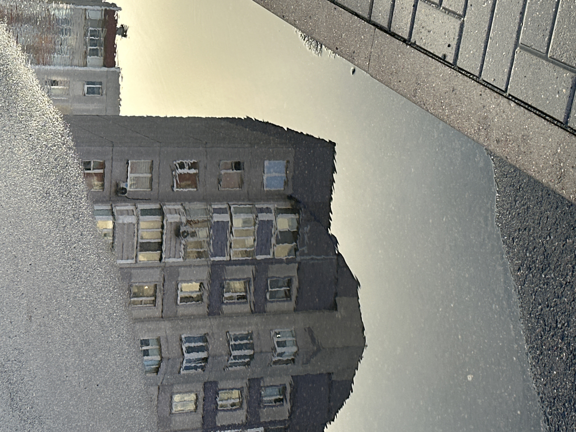The image shows a reflection of a multi-story apartment building in a puddle of water on a city street. The building appears to be made of concrete with numerous windows. The reflection is distorted by ripples on the water's surface. To the right of the puddle, a sidewalk paved with gray interlocking bricks is visible, bordered by a concrete curb. The scene suggests an urban environment in Minsk, Belarus, likely after rain. No people or vehicles are present in the frame. The lighting indicates it is likely daytime, possibly overcast or during twilight.