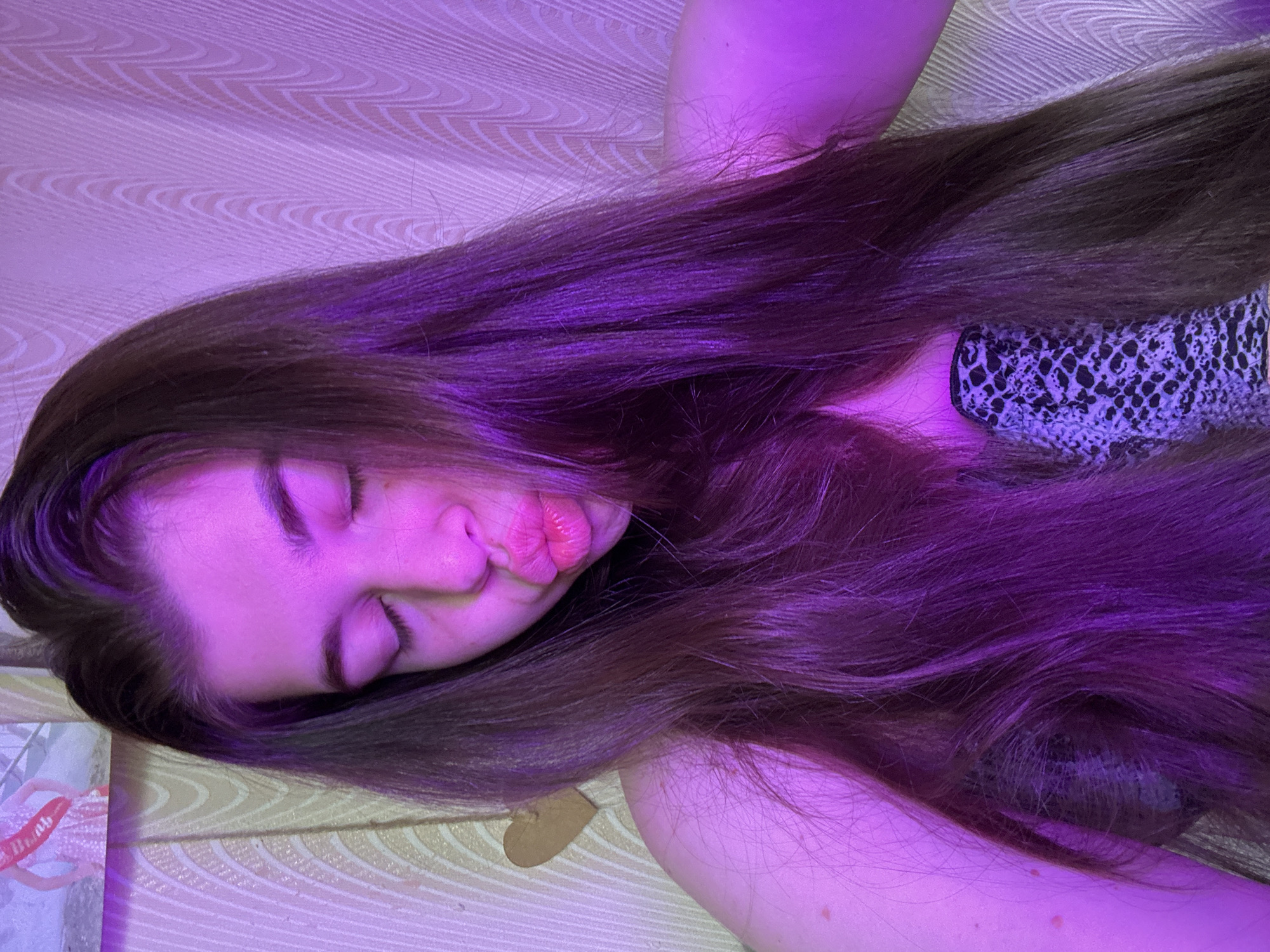 A young woman with long brown hair is lying down with her eyes closed and lips pursed. She is illuminated by a purple light. She is wearing a patterned top. The background appears to be a textured surface, possibly a wall or furniture. No information regarding the location in the city of Мещёры, Russia is evident.