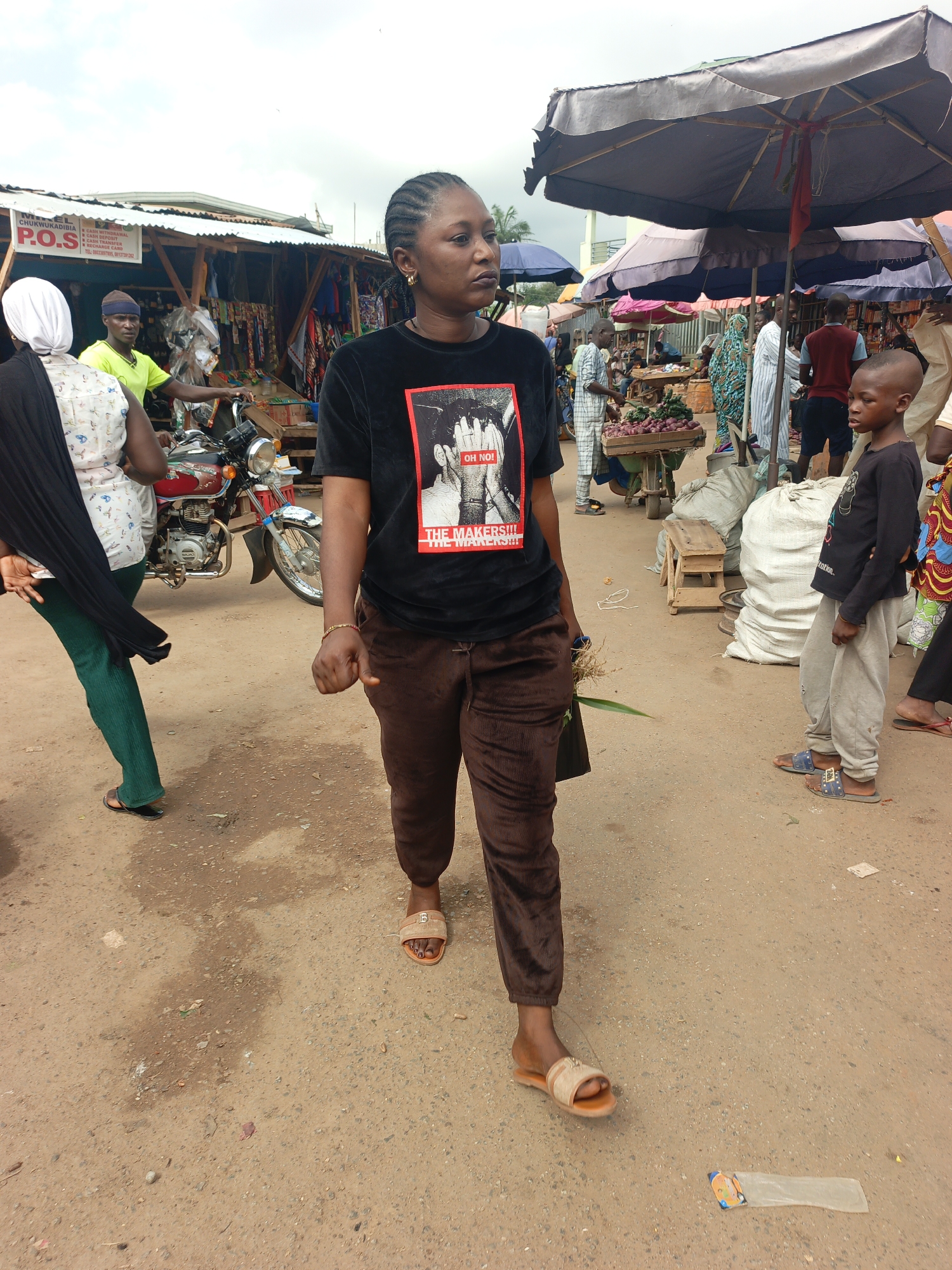 The image shows a woman walking through an outdoor marketplace in Akwanga, Nigeria. The ground is dirt and gravel. The woman is wearing a black t-shirt with an image on it, brown pants, and sandals. She is carrying something in her left hand. Various market stalls and vendors are visible in the background, with people browsing and buying goods. Motorbikes are parked nearby. A young boy is standing to the woman's right, wearing a long-sleeved shirt and sweatpants. Overhead are awnings providing shade to the market.