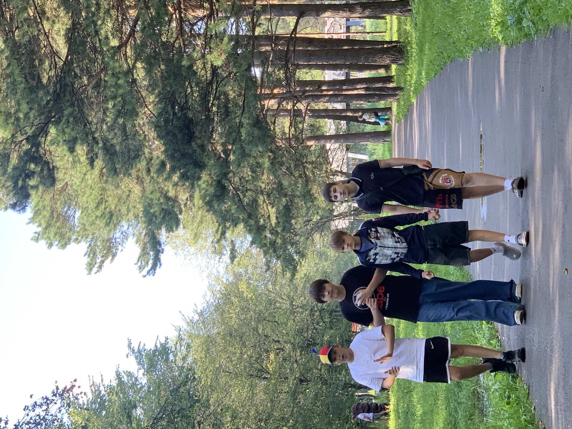 Four young males walk along a paved path in a park with tall pine trees and lush green grass. The person on the far left wears a white t-shirt and black shorts, with a colorful hat. The second person from the left wears a black t-shirt and blue jeans. The third person from the left wears a blue and white patterned jacket over a blue shirt and dark shorts. The person on the far right wears a black polo shirt and patterned shorts. In the background, other park-goers can be seen, including a person in a light blue shirt and dark pants. The environment suggests a warm, sunny day, typical of a summer in Prokopyevsk, Russia, a city known for its parks and natural beauty.