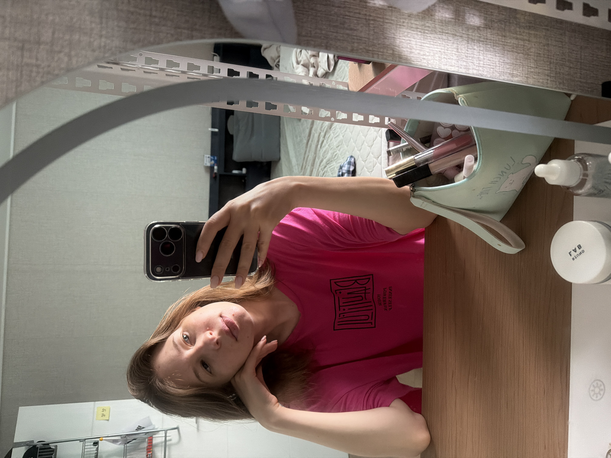 A young woman with blonde hair, wearing a bright pink t-shirt, is taking a mirror selfie in what appears to be her bedroom in Dongducheon-si, South Korea. She is leaning her head on her hand, looking directly at the camera. On the table in front of her, there is a makeup bag filled with cosmetics, a glass bottle with a dropper, and a round white container. In the background, a bed with patterned bedding and some shelves can be seen. The room is lit by natural light coming from a window, not visible in the frame. The overall atmosphere of the image is casual and relaxed.
