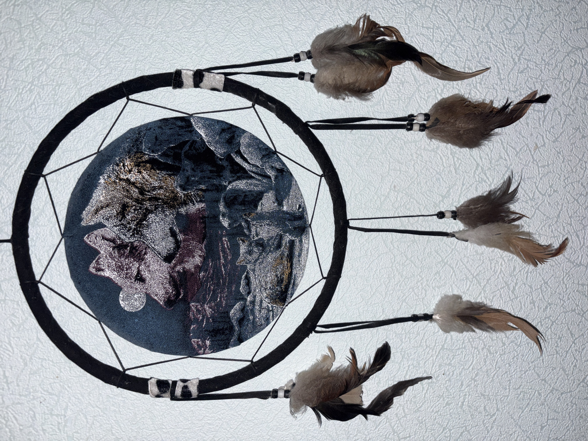 The image shows a dreamcatcher hanging against a textured, light-colored wall. The dreamcatcher has a black hoop with intricate webbing inside. The webbing features a circular tapestry depicting wolves in a rocky, moonlit landscape. Several black strings hang from the bottom of the hoop, adorned with feathers of various sizes and colors, some of which are fluffy and brown, while others are longer and darker. The background wall appears to be a wall in a room. No specific location in Sverdlovskiy, Russia is identifiable from the image.