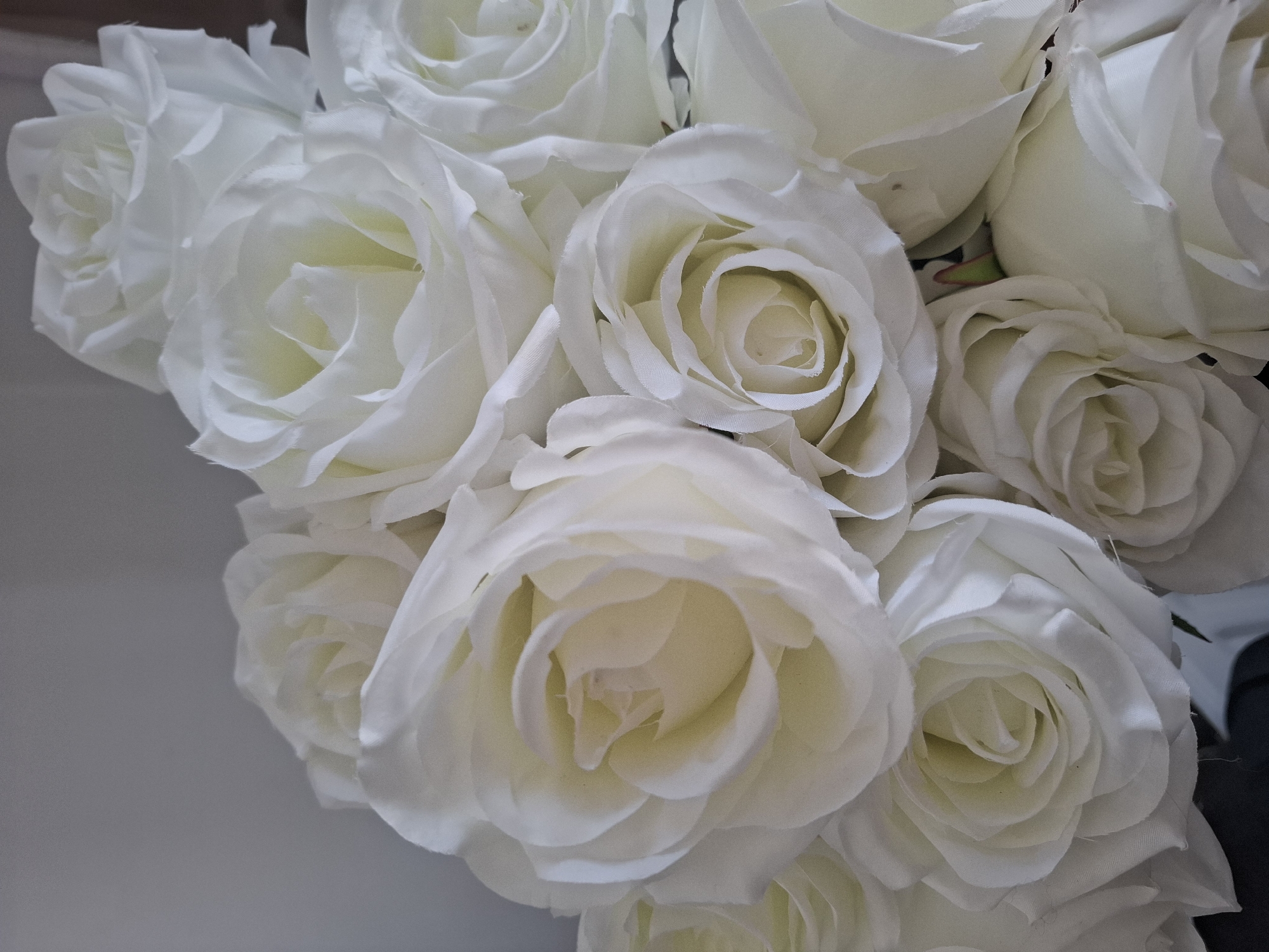 A close-up, high-angle shot shows a bouquet of white roses against a plain, light gray background. The roses are artificial, with fabric petals. They are arranged in a dense cluster, with some fully opened and others still partially closed. The lighting is soft and diffused, creating subtle shadows that highlight the texture of the petals. There are no people or other objects visible in the image. The scene appears to be indoors, possibly in a home or studio setting. No specific location in Montreuil, France can be identified from the image.