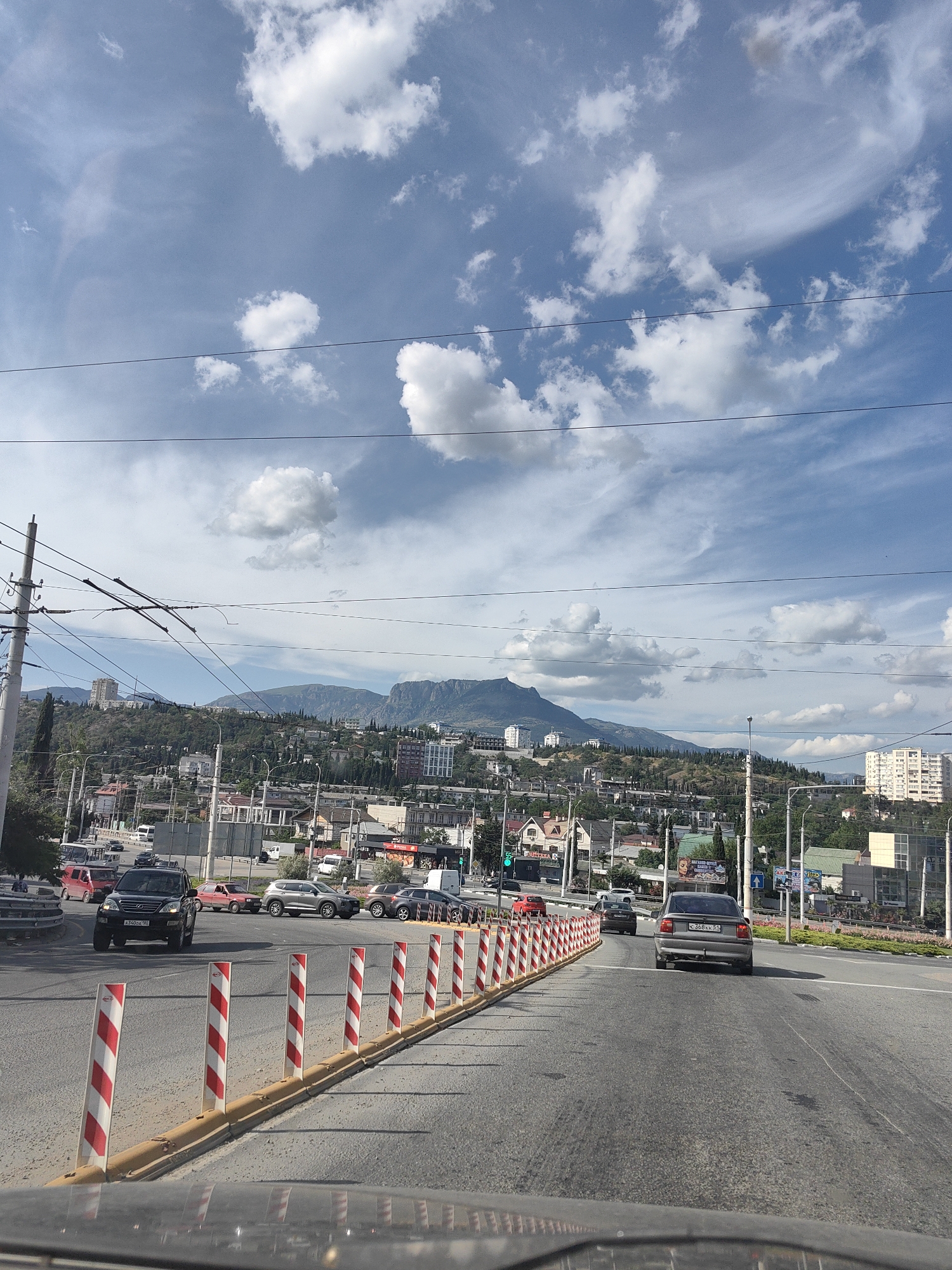 The image depicts a street scene in the city of Alushta, Ukraine, on a partly cloudy day. The perspective is from a vehicle moving on a road with multiple lanes. In the foreground, a row of red and white striped bollards line the right side of the road, separating lanes. Several cars are visible on the road, including a black SUV and a gray sedan. The background features a cityscape with buildings of varying heights, nestled on a hillside. Beyond the city, a large mountain range rises, partially obscured by clouds. Overhead, power lines crisscross the sky, supported by poles. The overall impression is that of a typical urban environment with a natural landscape as a backdrop.