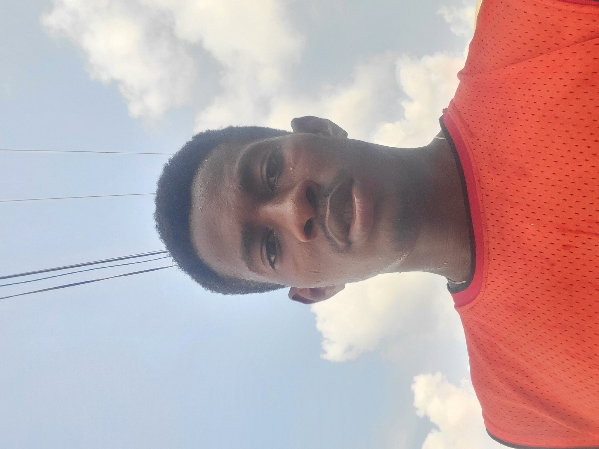 A medium shot, upward-angled selfie shows a young Black man in an orange mesh tank top, looking directly at the camera with a neutral expression. His forehead, cheeks, and neck appear shiny with sweat. The background is a bright, cloudy sky with three thin, dark power lines stretching diagonally across the upper portion of the frame. The image was taken outdoors, likely in Egbagu, Nigeria, based on the provided context.
