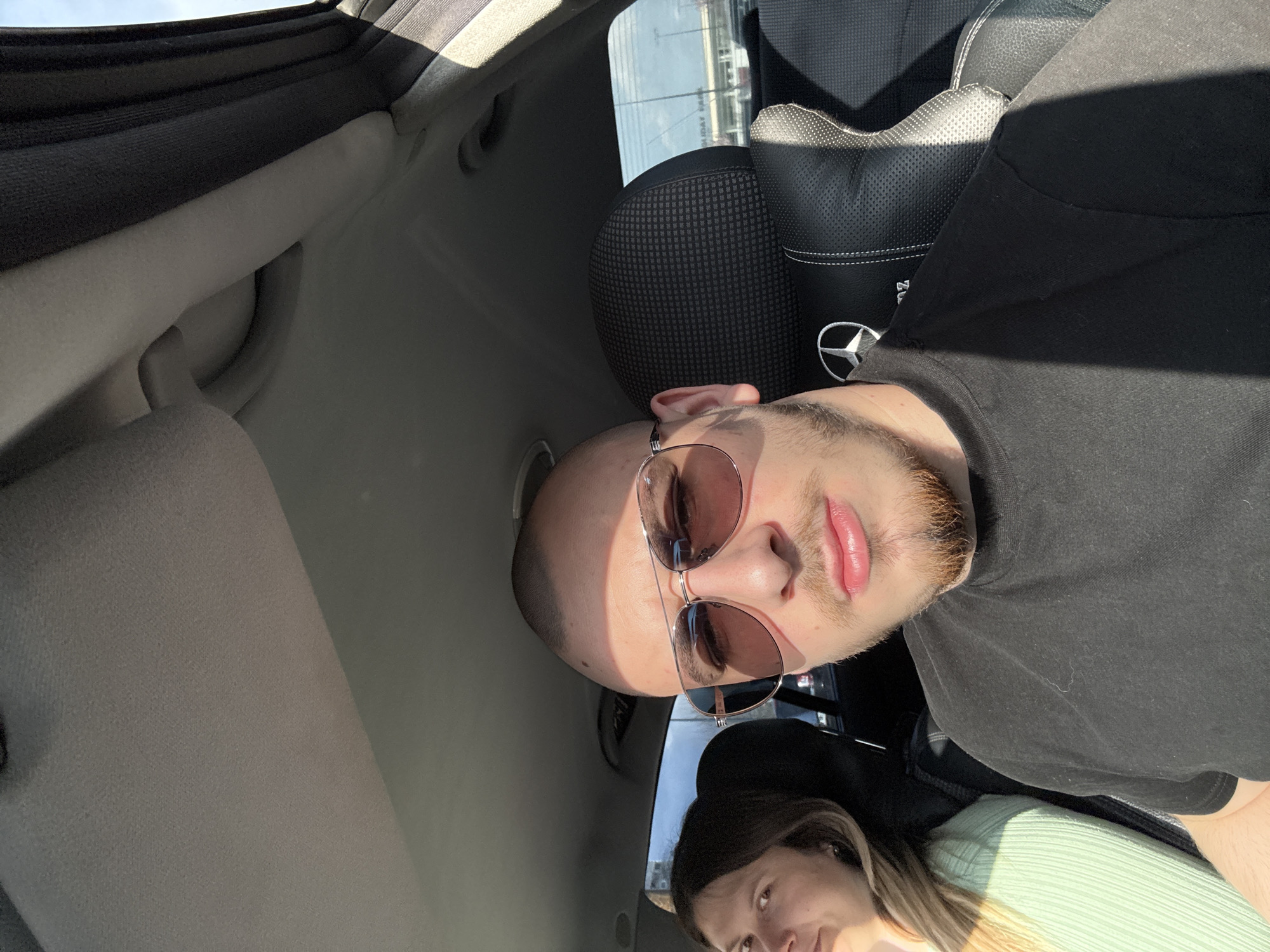 The image shows a selfie taken inside a car. A man wearing sunglasses and a black t-shirt is in the foreground. A woman with long brown hair and a light green sweater is visible in the lower portion of the frame. The interior of the car includes a headrest with a Mercedes-Benz logo. Through the car window, a building appears to be located in Stara Zagora, Bulgaria, although the exact location cannot be determined without further information.