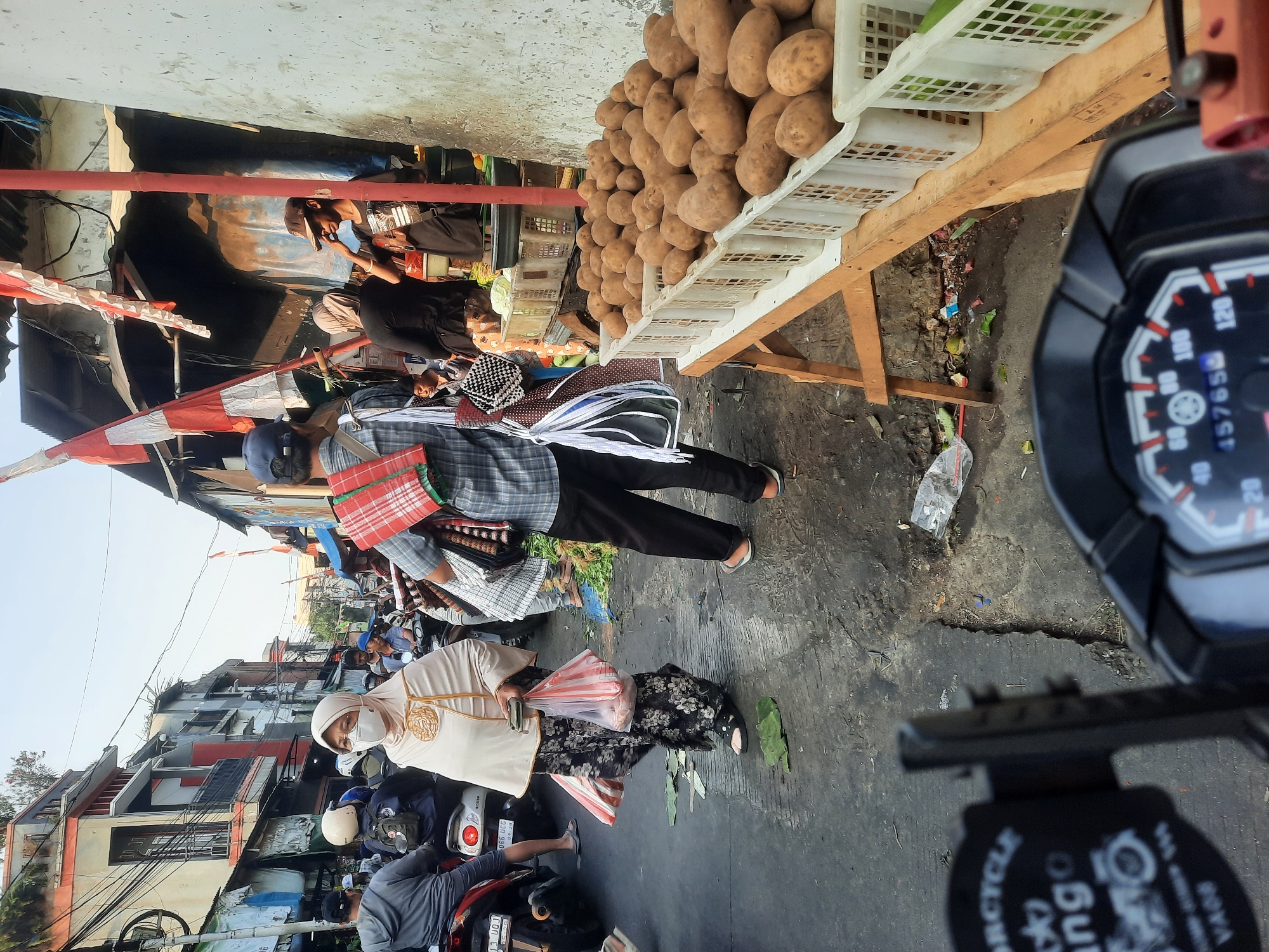 The image depicts a busy street market scene in North Jakarta, Indonesia. The foreground shows the dashboard of a motorcycle, suggesting the photographer is on a motorbike. The middle ground features a variety of people walking and riding motorcycles, with some wearing masks. A woman in a beige hijab and floral skirt is visible, carrying plastic bags. To the right, a stall displays a large quantity of potatoes in white plastic crates on a wooden table. Behind the stall, a building with a rough, textured wall is visible, and further back, a row of buildings with various architectural styles lines the street. Indonesian flags are displayed on some of the structures. The ground is paved and appears wet in some areas, with scattered debris. The overall atmosphere suggests a bustling local marketplace.