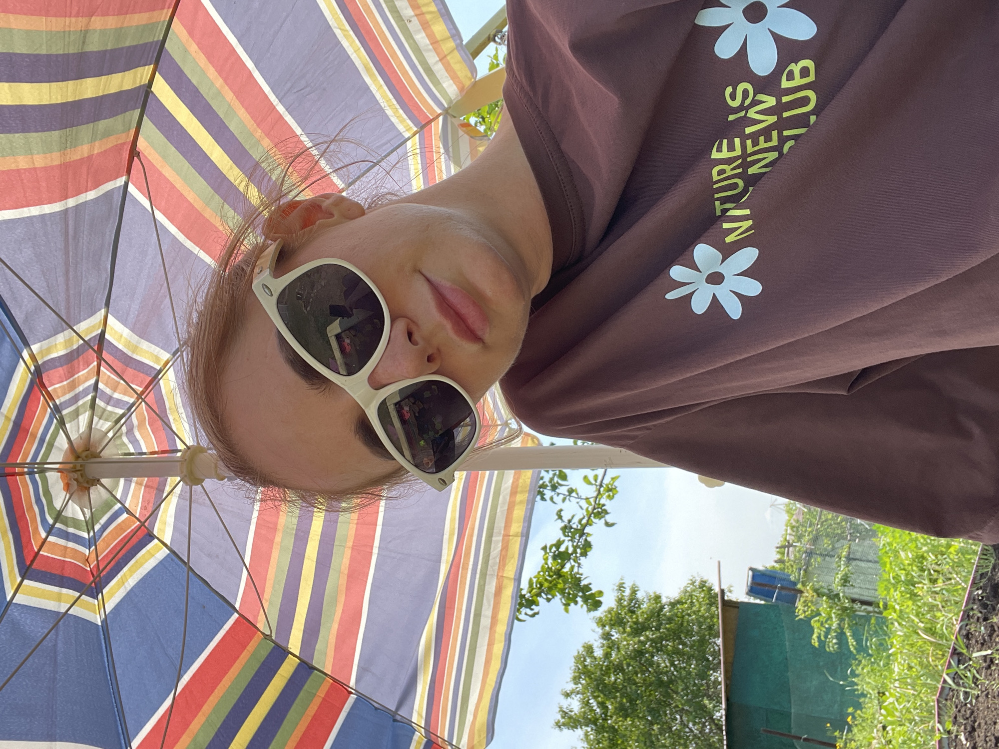 A low-angle, rotated perspective image shows a person in sunglasses and a brown long-sleeve shirt with white flower graphics and the text "NATURE IS NEW CLUB." The person is standing under a colorful striped umbrella. In the background, partially obscured by the umbrella and the person, is a scene of greenery, possibly a yard or garden, with trees and a fence. The presence of a building with a blue roof is also evident. The location, Yasnoe, Russia, cannot be verified solely from the image content.