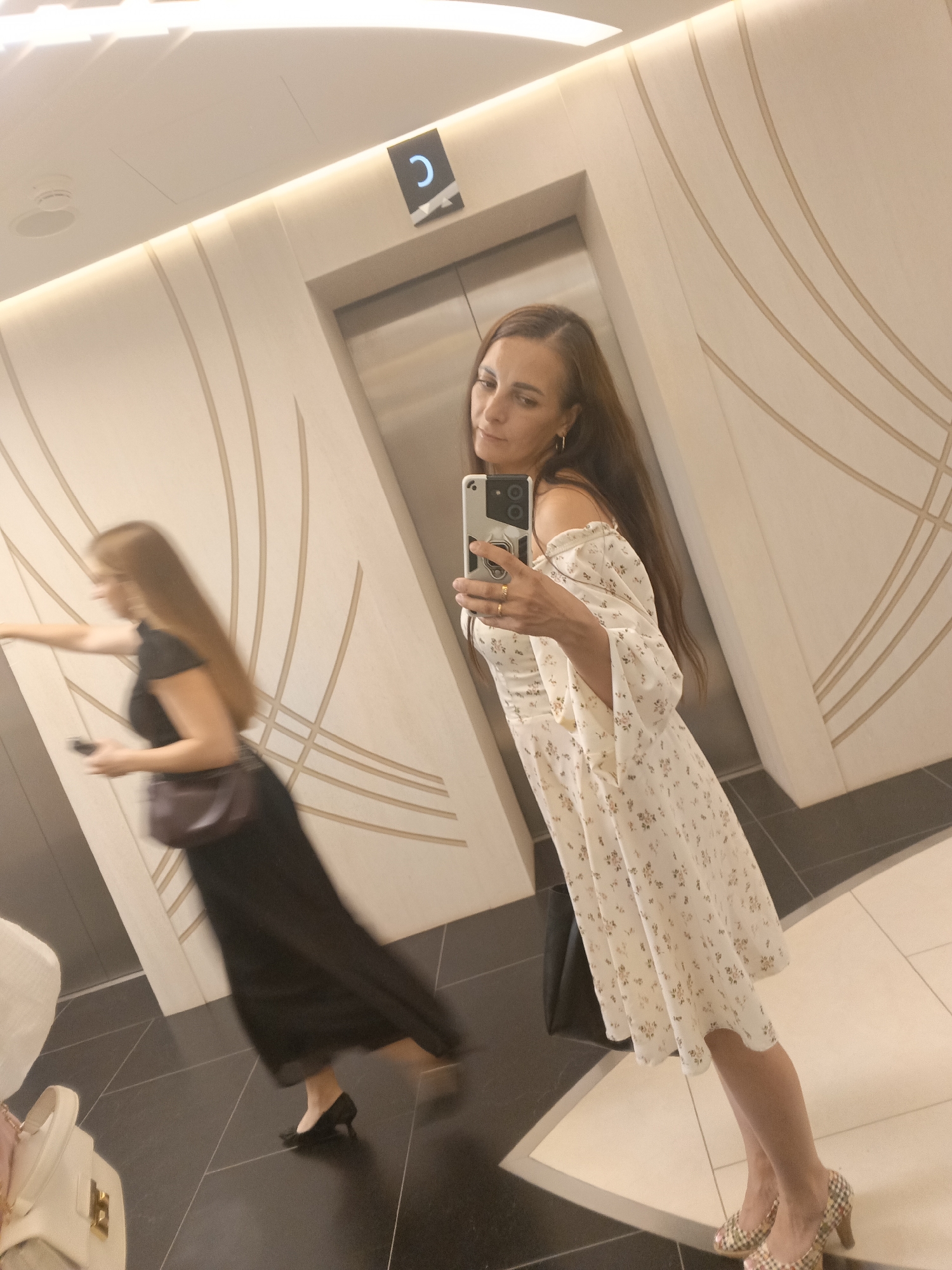 In a dimly lit, modern hallway, two women are reflected in a large mirror. The woman on the right, in the foreground, is wearing a white off-the-shoulder dress with a floral pattern and leopard-print heels. She is holding a smartphone, taking a selfie. Her hair is long and brown. The woman on the left, in the background, is blurred due to motion, wearing a black dress and a dark cross-body bag. She is walking away from the viewer, holding a phone in her left hand. The hallway walls are decorated with a pattern of curved lines, and an elevator door is visible behind the women. The floor is tiled with black and white squares. The scene appears to be in Moscow, Russia, judging by the Cyrillic "C" sign above the elevator, which likely indicates the floor or a section of the building.
