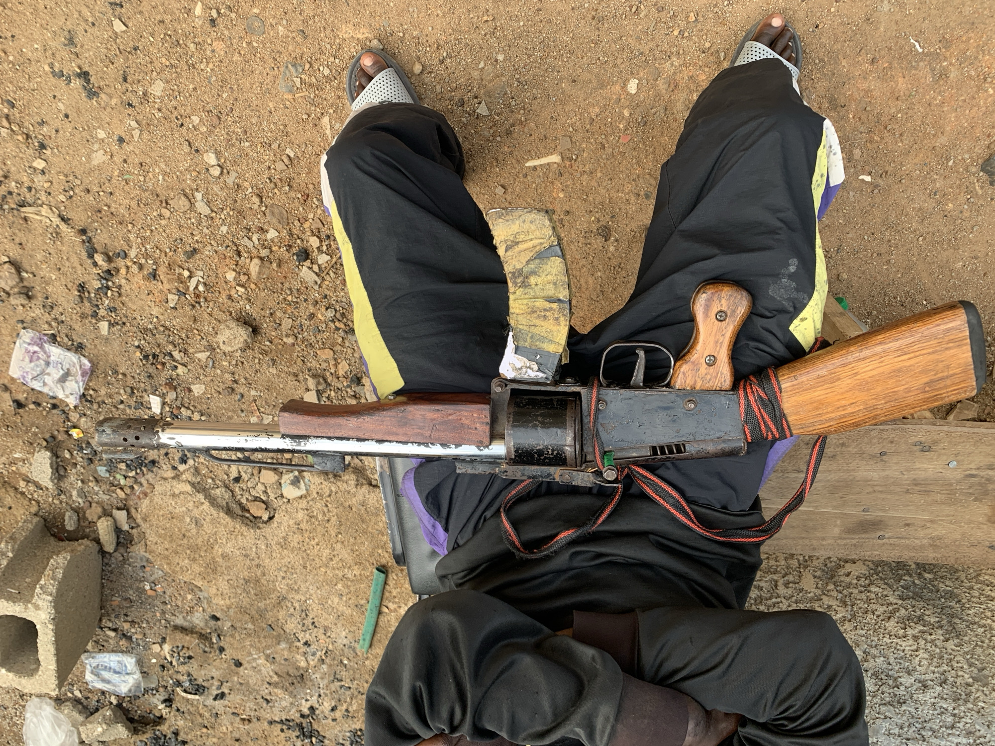 The image depicts a person sitting with an AK-47 assault rifle resting on their lap. The person is wearing dark-colored pants with yellow and purple accents, and sandals. The rifle has a wooden stock and grip, with a red and black sling. The background consists of a dirt ground with scattered debris and a concrete block. The location is likely New Stadium, Nigeria, given the context.