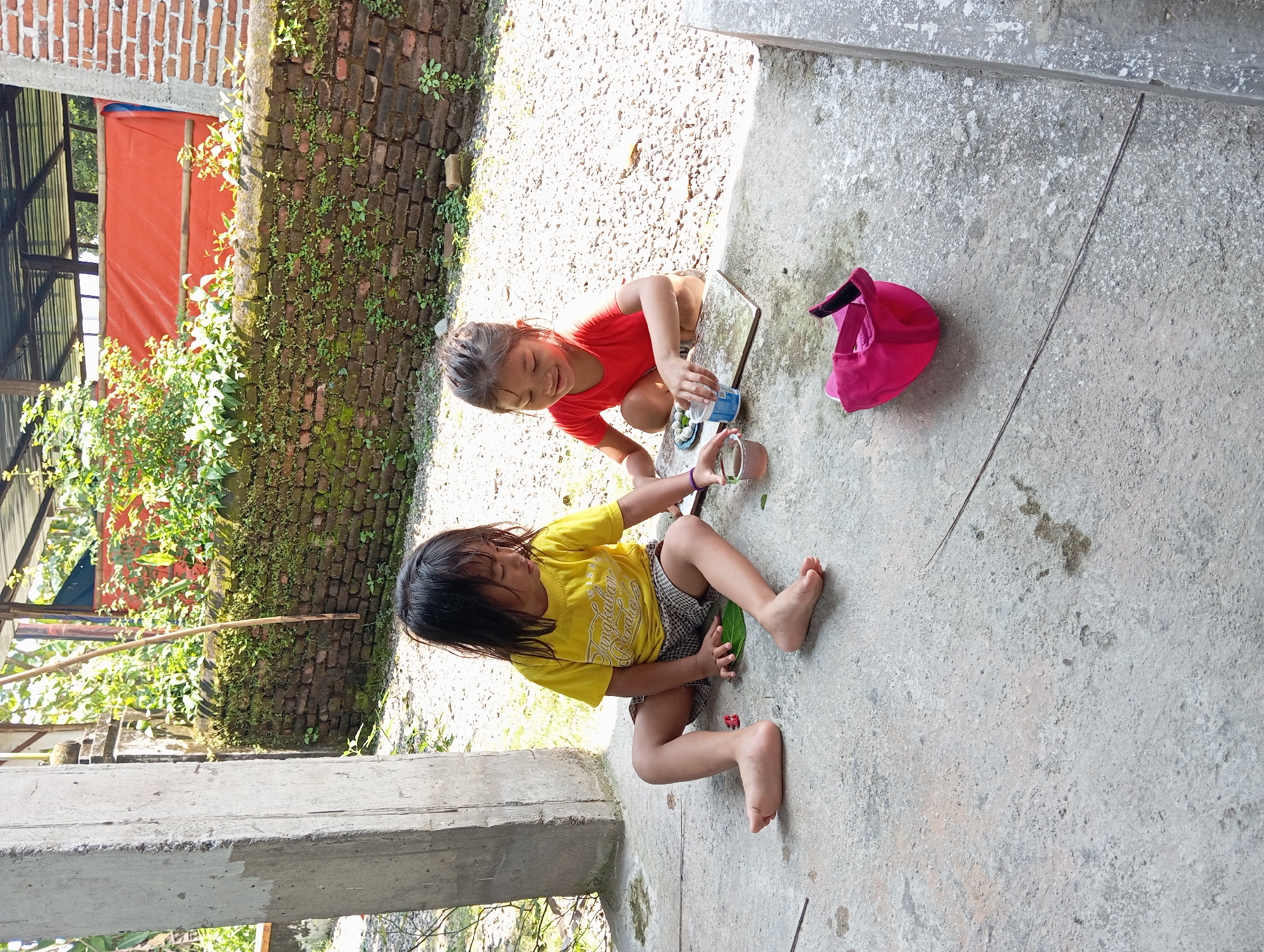 Two children are playing outdoors on a concrete surface. One child, wearing a red shirt, kneels while interacting with a small blue and white plastic container and a clear glass. The second child, wearing a yellow t-shirt, sits with their legs extended. A pink hat sits on the concrete beside the kneeling child. A brick wall with moss growth is visible in the background, along with greenery and a structure covered in orange tarp. The photo was taken in Baron, Indonesia.