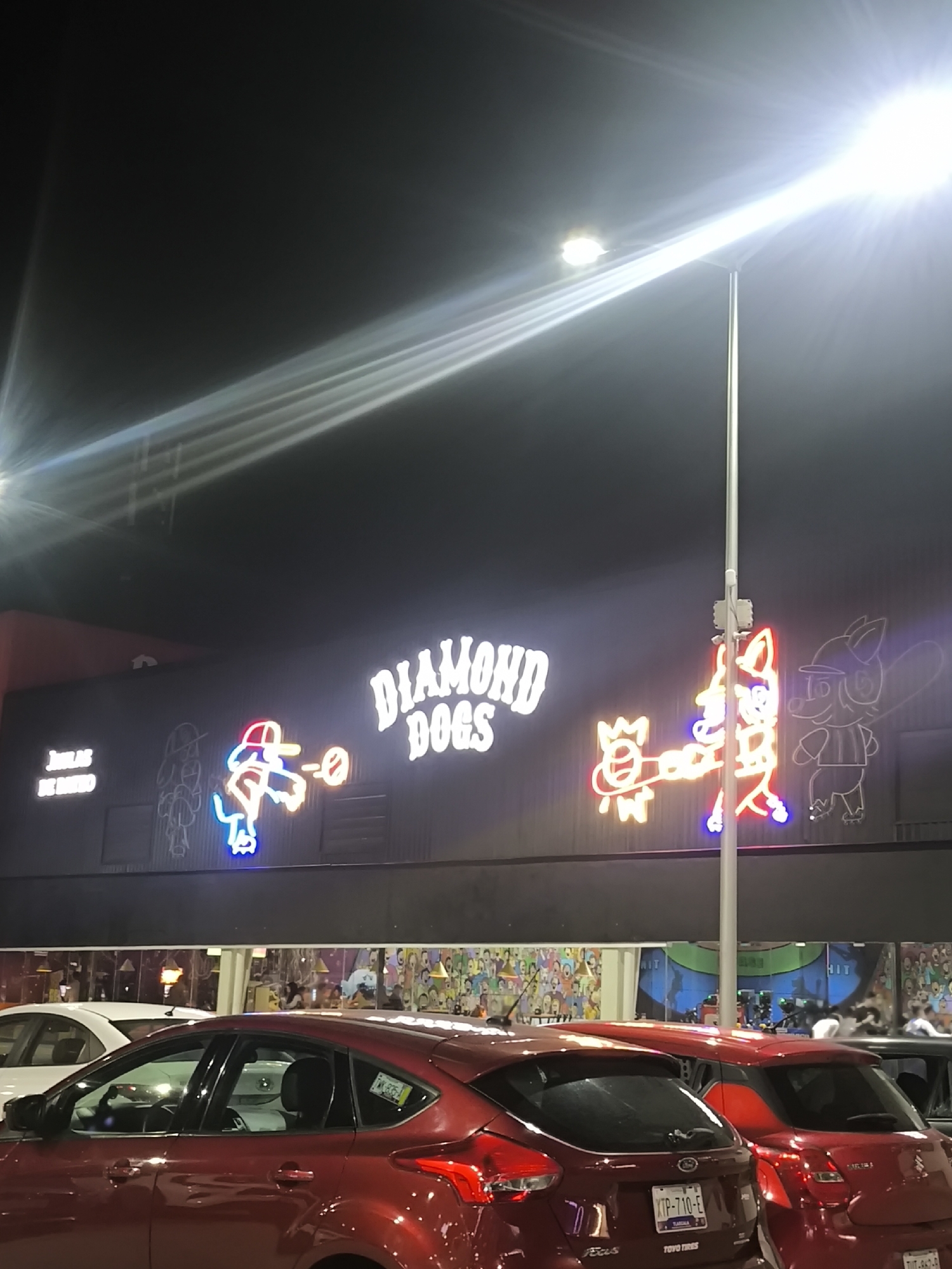 At night in San Andrés Cholula, Mexico, a row of cars is parked in front of a building advertising "Diamond Dogs." The cars include a red Ford Focus with the license plate "KTP-710-E." The building features neon signs of baseball-themed dog characters and the business name. A mural with numerous cartoon faces is visible behind the parked cars. A street light illuminates the scene.