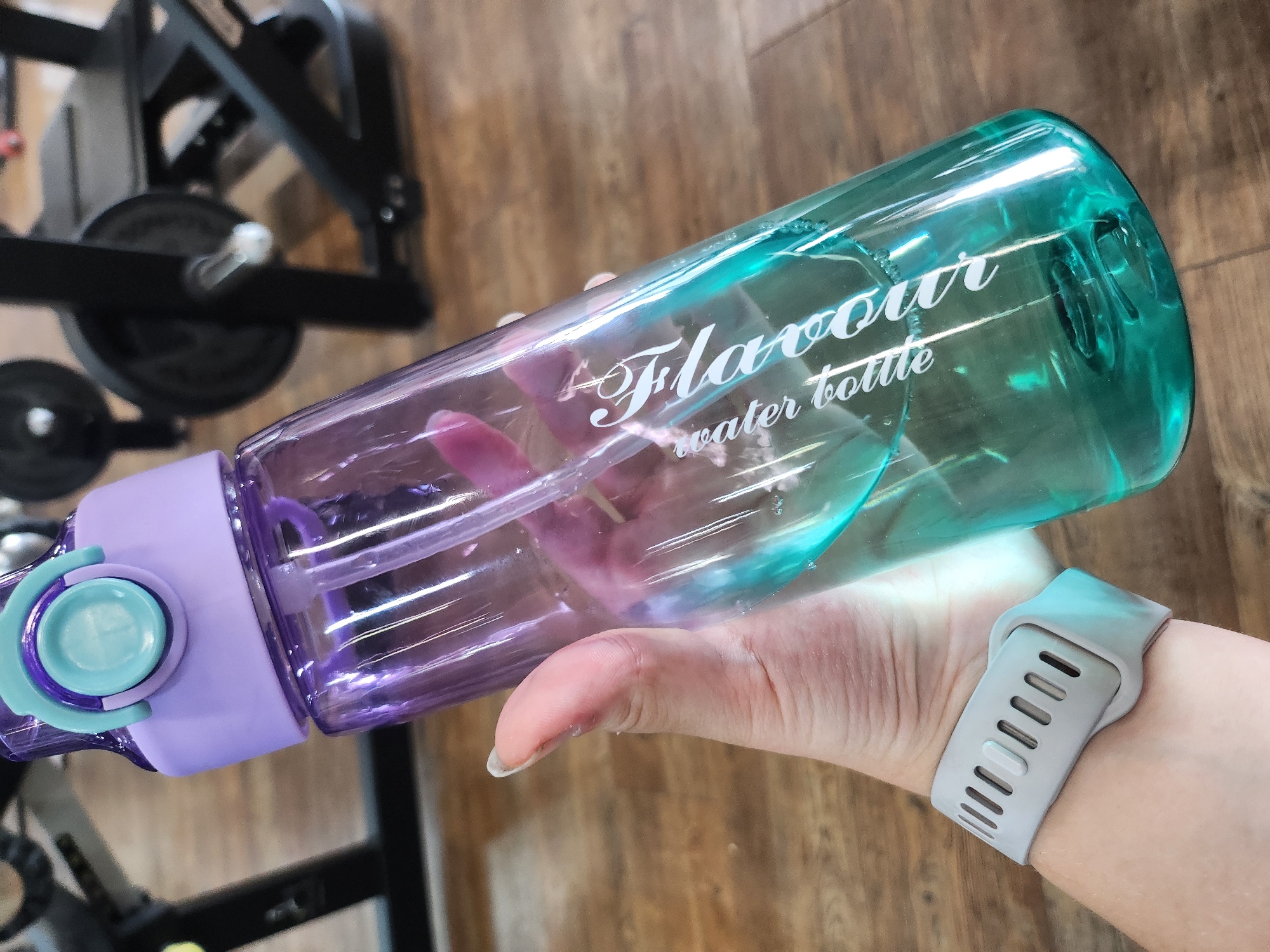 A person's hand holds a two-tone water bottle in an indoor setting in Chaikovsky, Russia. The transparent bottle features a gradient color, transitioning from light purple at the bottom to a teal-green hue towards the top. Its lid is light purple with a teal-colored push-button or clasp mechanism. White script text on the bottle reads "Flavour water bottle." A light grey smartwatch or fitness tracker with a perforated strap is visible on the person's wrist. The background is blurred but shows elements of gym equipment, including black metal frames and circular weights. The floor appears to be constructed of light brown wooden planks. The scene captures a moment, likely during or after a workout, in a gym or fitness center.