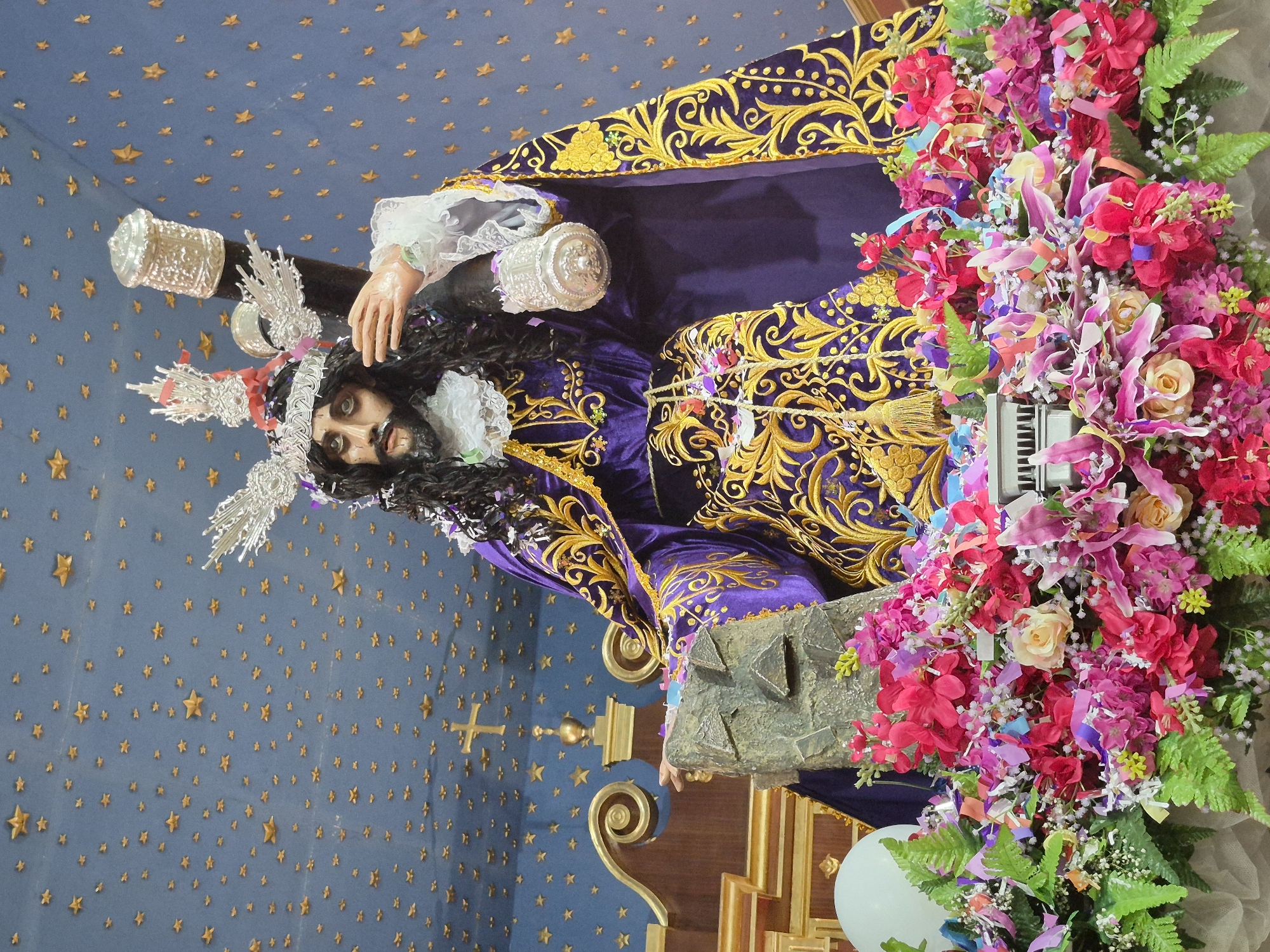 A full shot of a statue of Jesus Christ carrying a cross is on display in front of an altar in the Church of Pozo Almonte, Chile. The statue is adorned with a purple robe embroidered with gold patterns, a silver crown with red accents, and a silver halo. Jesus is depicted with long dark hair and a beard, his eyes are open and looking forward. He is holding the cross on his right shoulder, with his left hand resting on a stone slab. The background is a blue wall decorated with numerous small gold stars, and a gold cross emblem is visible on the wall. The statue is surrounded by an abundance of colorful flowers and confetti, forming a vibrant floral arrangement at its base. The altar itself is visible behind the statue, featuring wooden textures and gold embellishments.