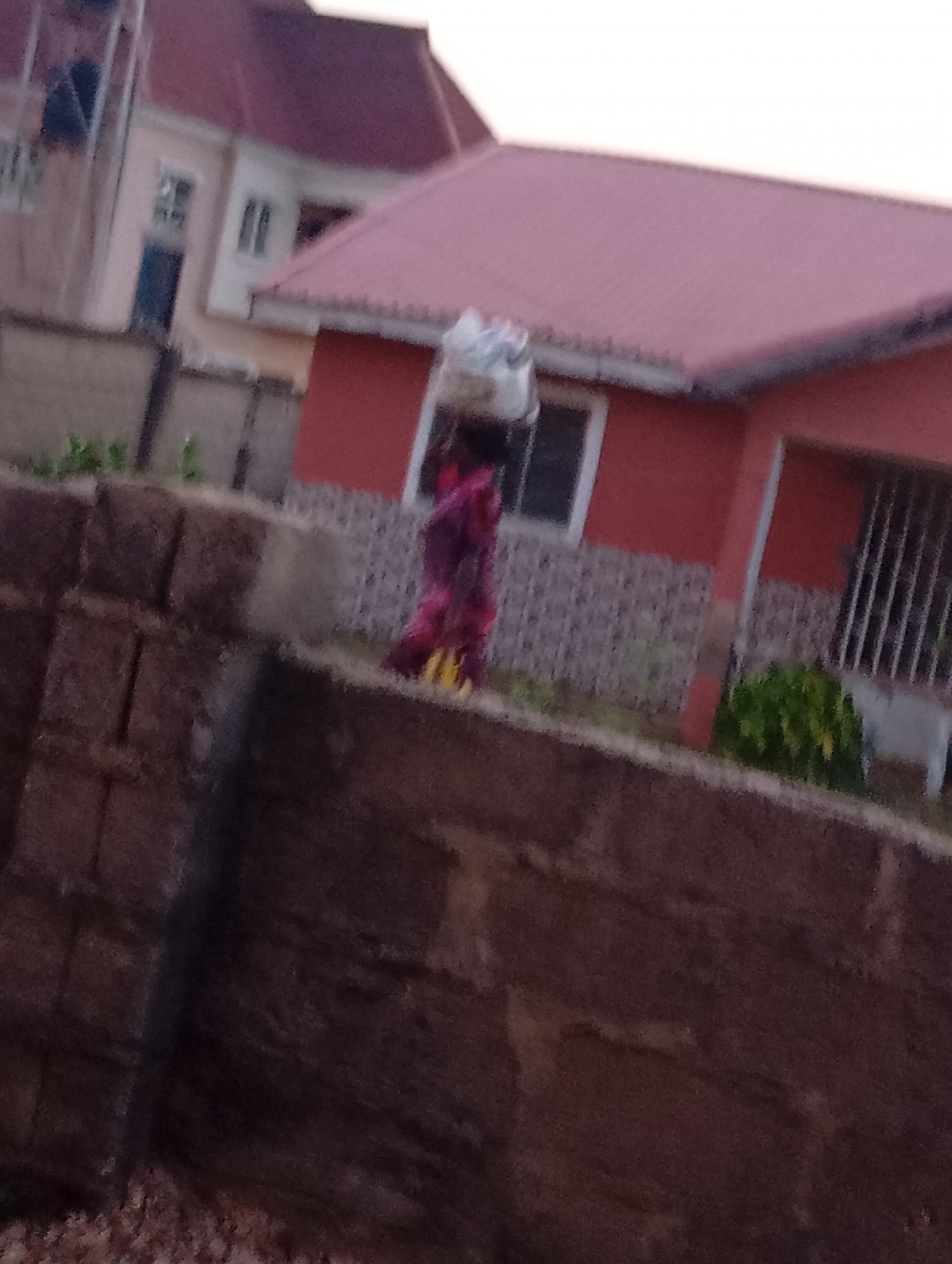 The image captures a residential scene in Unknown, Unknown. A person wearing a pink patterned outfit and carrying a large white bundle on their head is seen in the middle ground. Behind them is a red house with a reddish-brown roof. Part of another house in lighter shades is visible in the background. A stone wall is prominent in the foreground. The lighting suggests it might be evening or dusk.