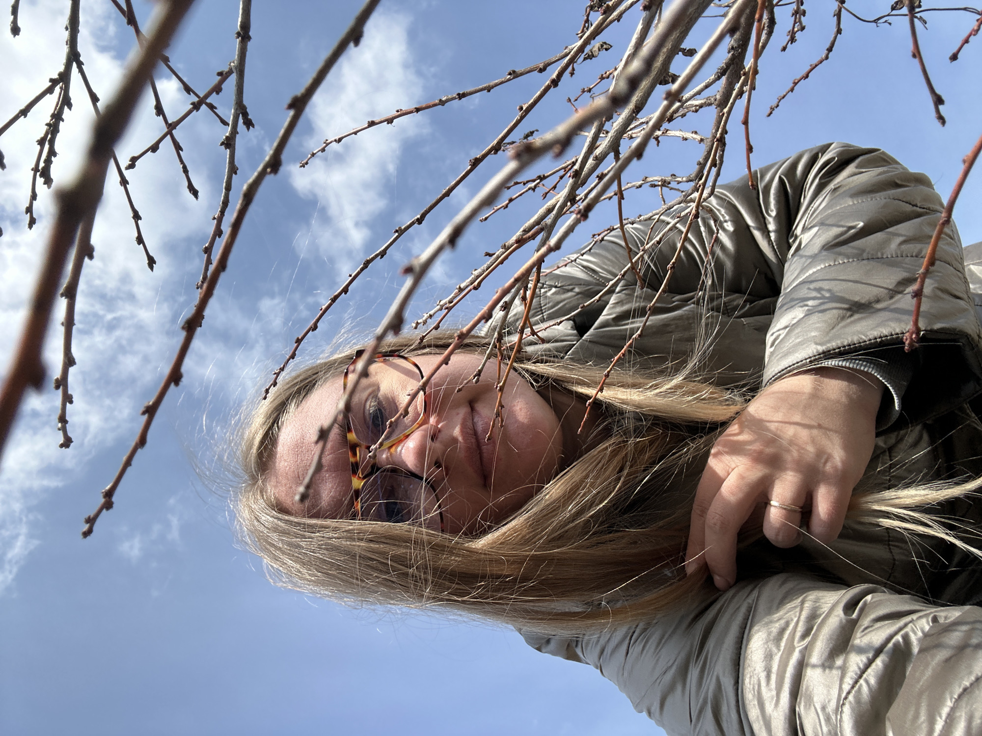 A low-angle shot taken outdoors in Уральское, Russia, shows a woman with long blonde hair and tortoiseshell glasses. She's wearing a metallic-colored puffy jacket. Her hand, with a gold ring on one of the fingers, rests on her hair. Bare tree branches frame the top of the shot against a light blue sky with scattered clouds.
