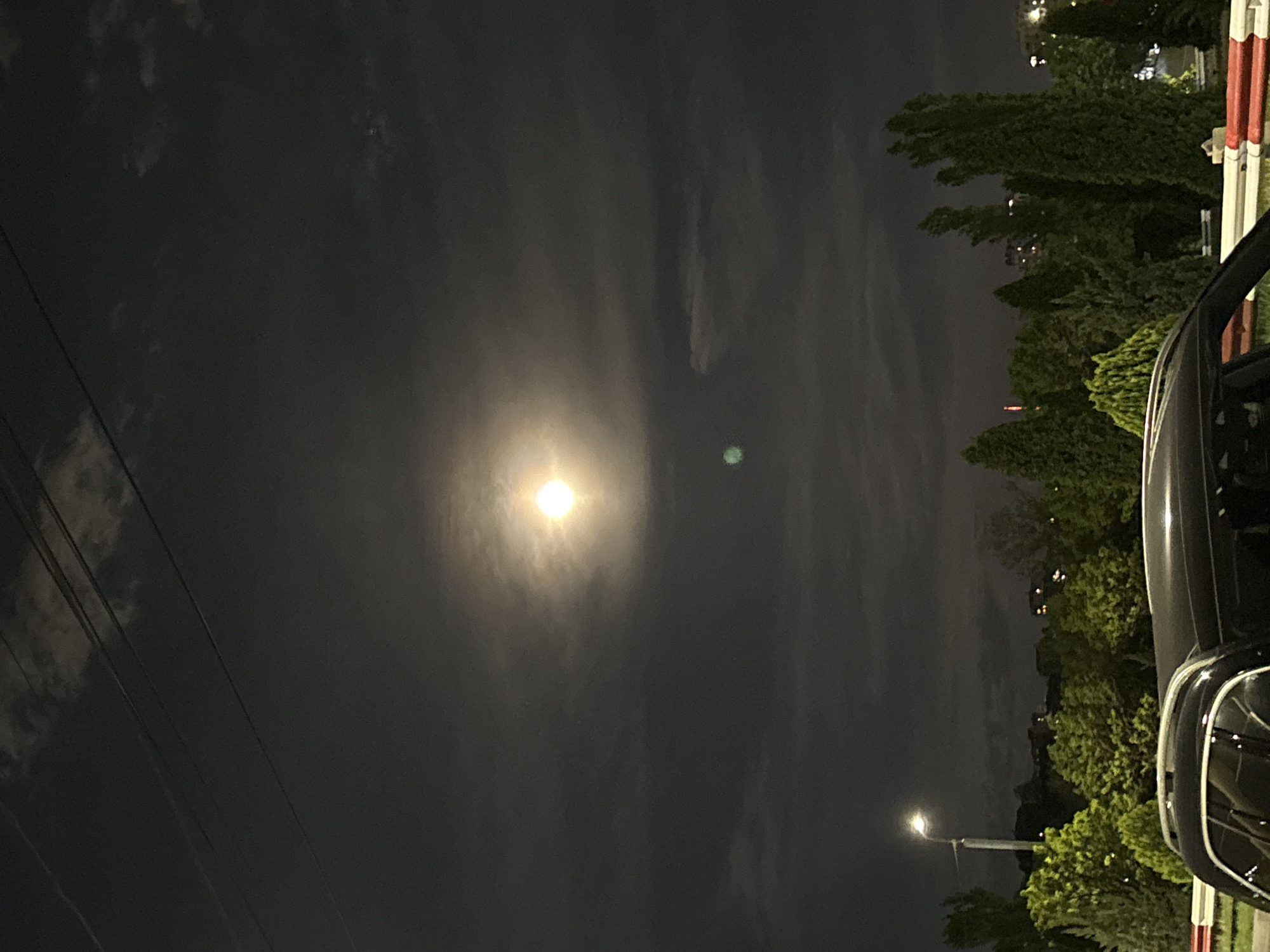 The image captures a serene night scene, dominated by a bright, luminous moon shining through a hazy, dark sky. The moon is positioned centrally, emitting a soft glow that subtly illuminates the surrounding wispy clouds.
In the upper left portion of the frame, several dark utility lines diagonally cross the dark sky. Along the right side of the image, a dense line of green trees stands against the night, with some distant, faint lights visible through their foliage, suggesting nearby buildings or an urban environment.
On the far right, a partial view of a dark-colored vehicle, likely a car, is visible, showing its front quarter and a portion of its side mirror. Directly behind the car, a red and white striped barrier or pole can be discerned. In the lower right corner, a single streetlamp emits a warm, yellowish light, illuminating a small area beneath it and the nearby tree branches. A noticeable green lens flare appears just to the right of the moon.
The scene conveys a quiet, late-night atmosphere. There are no visible people or discernible activity taking place. The time of day is clearly night, characterized by the dark sky, bright moon, and artificial light sources. No text is visible in the image.