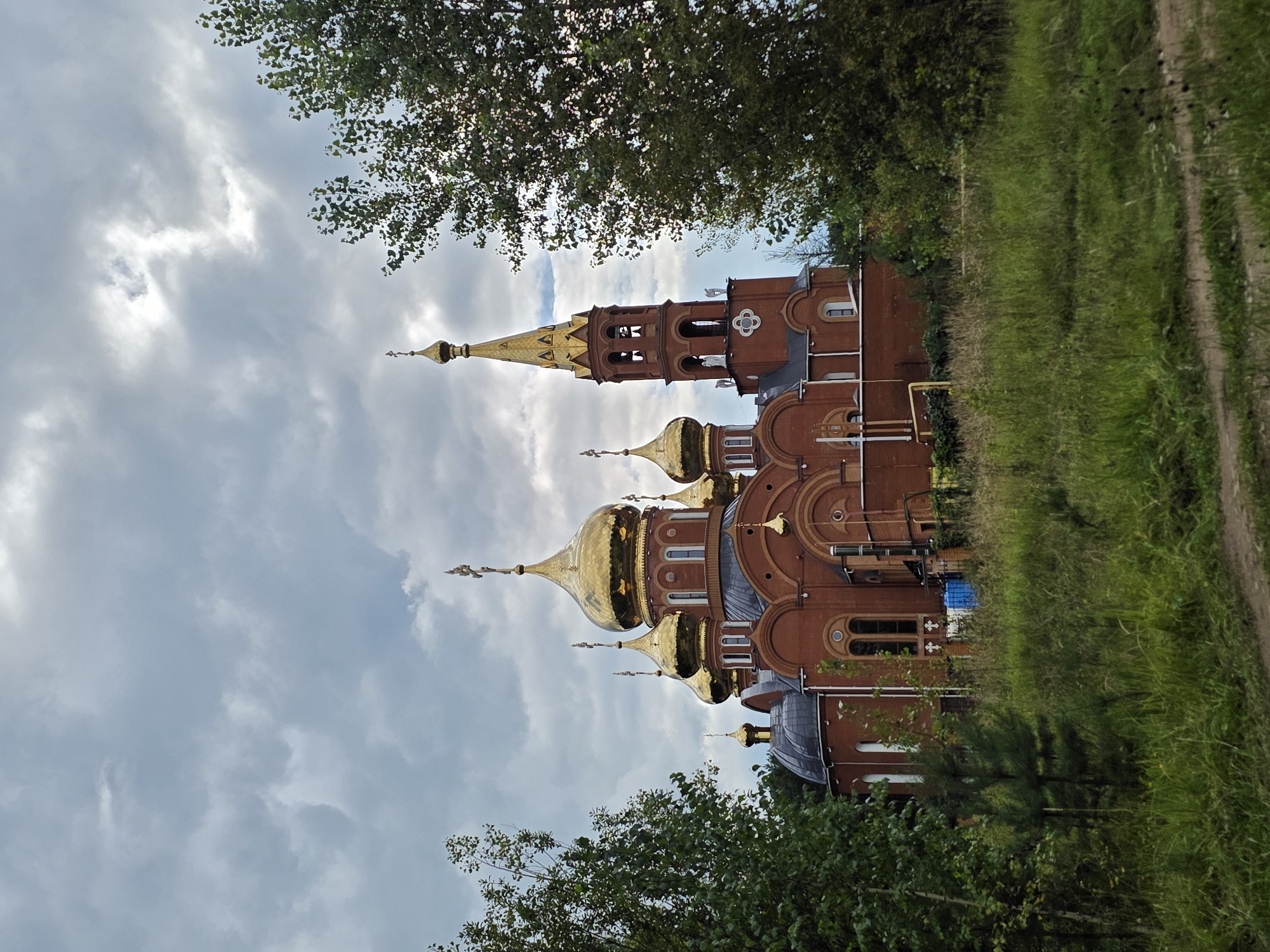 The image shows a brick Orthodox church with golden domes, located in Chaikovsky, Russia. The church has multiple domes, with the tallest being a bell tower topped with a golden spire. The building is surrounded by green trees and overgrown grass, suggesting a somewhat rural or less developed area. The sky is cloudy and overcast, with patches of lighter clouds indicating potential sunlight breaking through. There are no people or animals visible in the image. The overall scene depicts a religious structure in a natural setting under a dramatic sky.