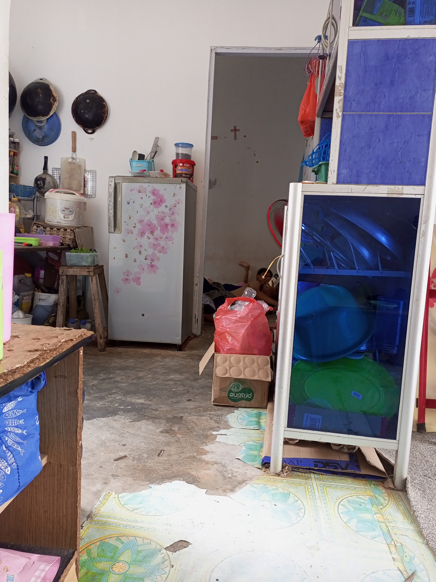 The image depicts a cluttered indoor space, likely a kitchen or living area, in Batam, Indonesia. The room is dimly lit, with concrete floors and white walls. Various household items are scattered throughout, including a refrigerator adorned with floral decals, a rice cooker, and stacks of boxes. A person, partially visible, is lying on the floor in the background, seemingly engrossed in a mobile phone. A metal and blue-paneled cabinet stands prominently in the foreground, filled with dishes and bowls. The overall scene suggests a lived-in environment with a sense of disarray.