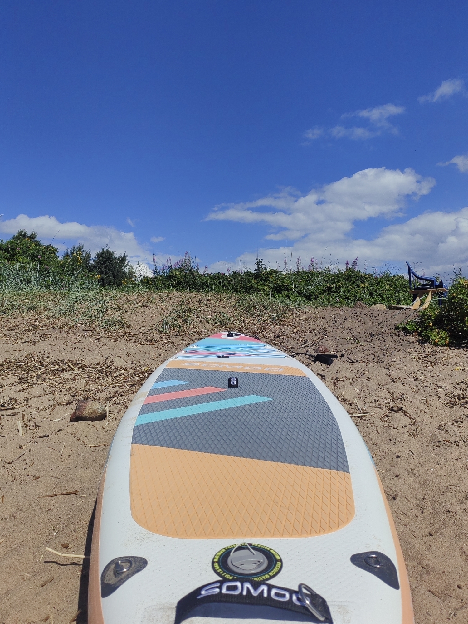 A stand-up paddleboard rests on a sandy beach, its nose pointing towards a lush green embankment under a bright blue sky dotted with white clouds. The paddleboard features a colorful design with orange, grey, and teal geometric patterns on its surface. Behind the beach, the embankment is covered in tall grasses and scattered pink flowers. In the background, a blue plastic chair and a yellow object are visible amidst the foliage, and beyond that, a body of water is suggested by the horizon. The scene is consistent with a location along the Neva River or the Gulf of Finland in Saint Petersburg, Russia, where such recreational activities are common during warmer months.