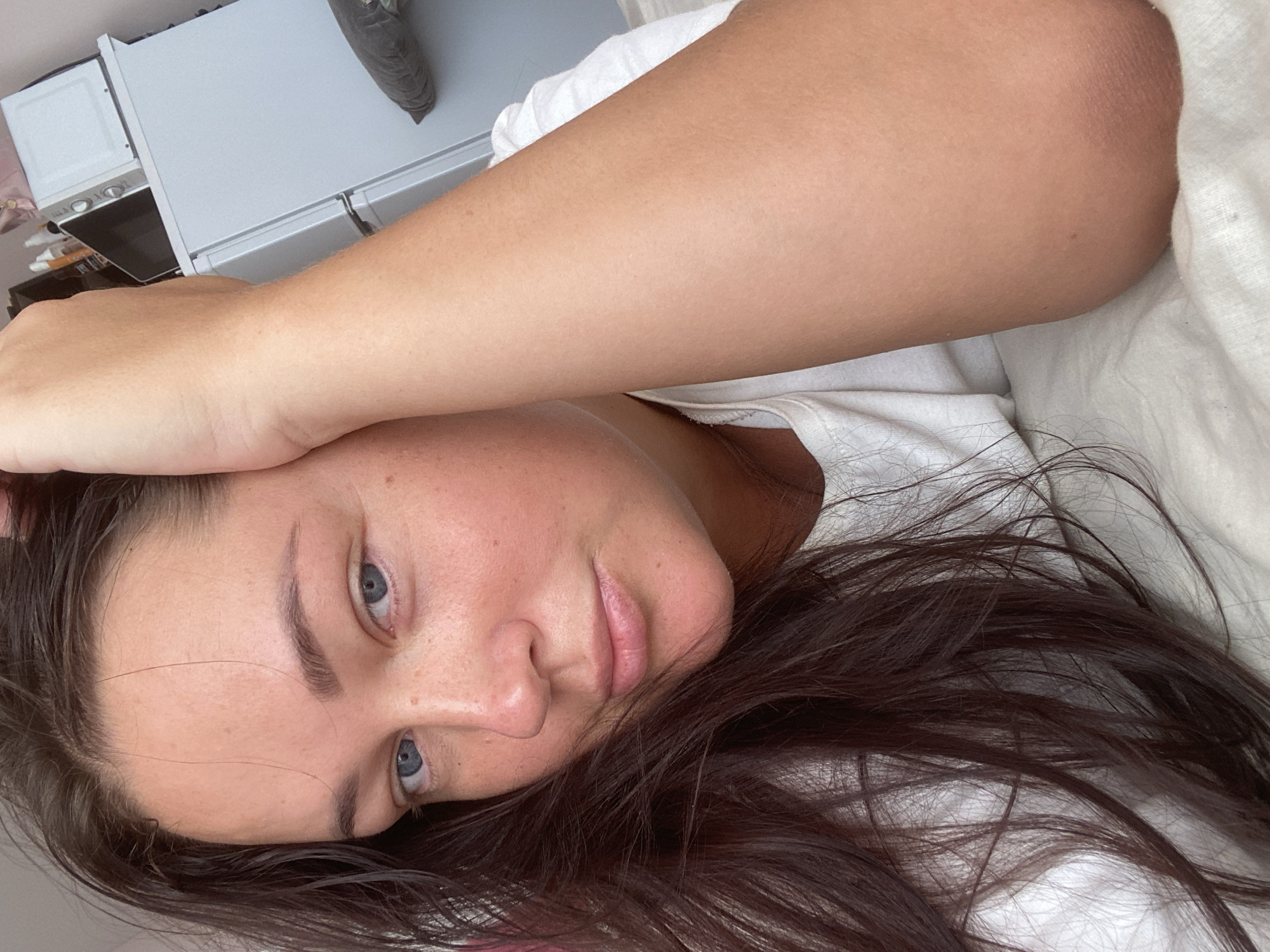 A young woman with brown hair and blue eyes is lying on a bed, with her right arm resting on her head. She is wearing a white t-shirt. The background shows a white refrigerator with a microwave on top. The scene appears to be indoors. The location of Sochi, Russia is not depicted in the image.