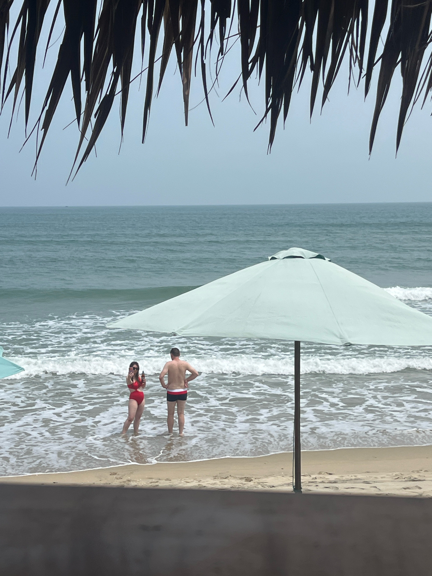 A full shot captures a beach scene in Hoi An, Vietnam, with a focus on two individuals standing in the shallow ocean water, partially obscured by a large, light blue beach umbrella. In the foreground, dry palm fronds hang down, creating a natural frame. The ocean stretches out to the horizon under an overcast sky. To the left, a portion of another beach umbrella is visible.
The woman on the left is wearing a red bikini and holding a bottle. She appears to be taking a photo with her phone. The man on the right is wearing dark swim trunks with a white waistband and has his hands on his hips. Both individuals are facing the ocean. The waves are breaking gently on the sandy shore. The overall mood is relaxed and leisurely.