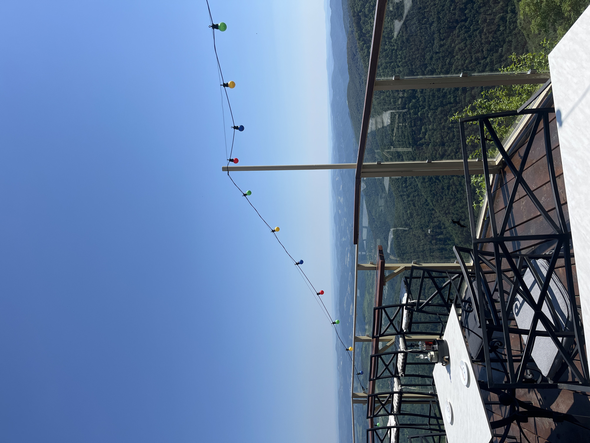 This is a landscape shot of a patio dining area with a panoramic view of rolling hills and a clear blue sky. The patio is furnished with black metal tables and chairs, some with white cushions. A string of colorful lights is strung overhead. The railing of the patio is made of glass and metal, offering an unobstructed view of the scenery. In the foreground, a white table is set with two white plates. In the background, a vast expanse of green forest stretches out to the horizon, dotted with faint outlines of buildings and roads. The lighting suggests it is daytime, likely late morning or early afternoon. No people are visible in the image.