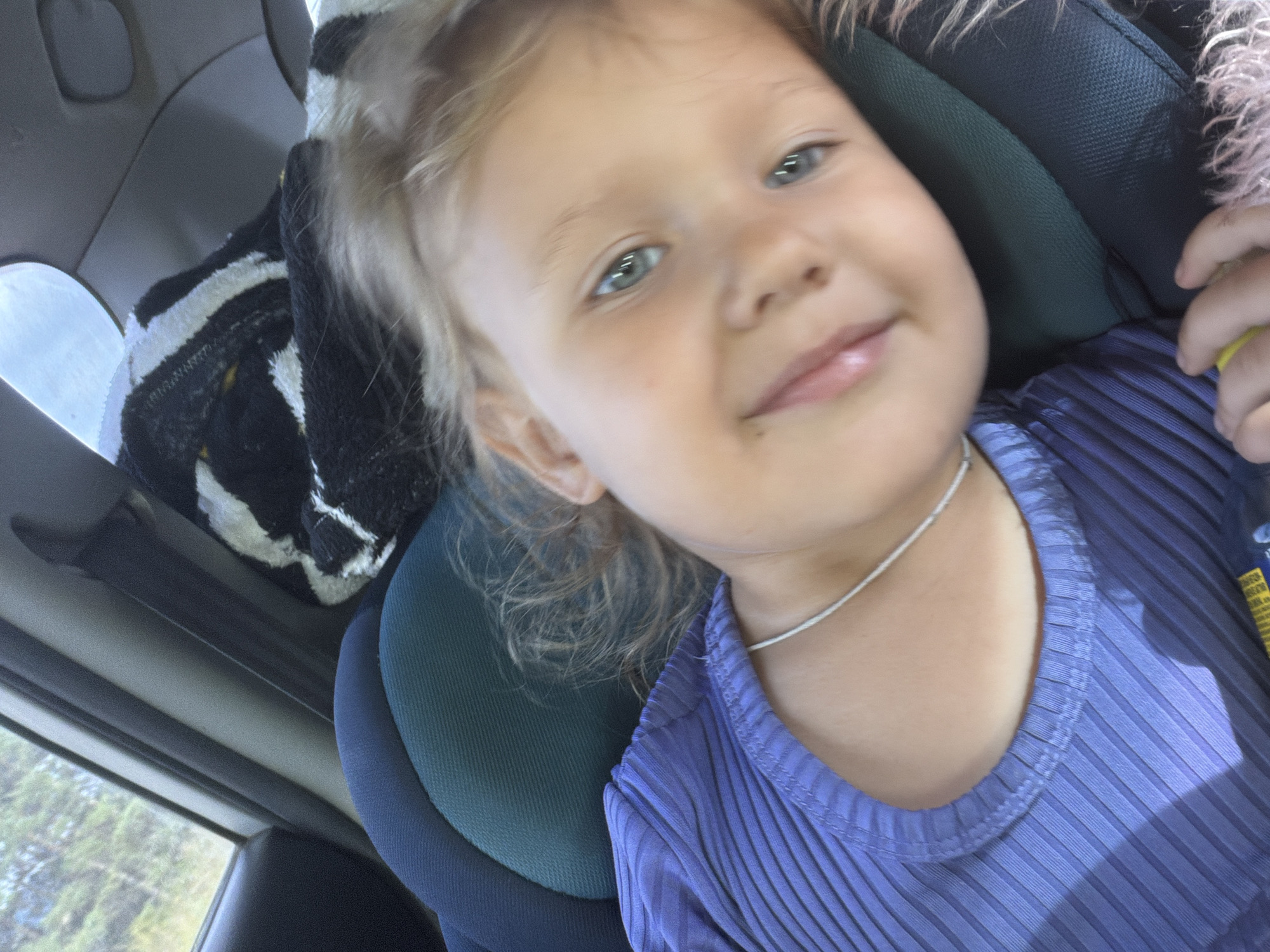 A young Caucasian child with blonde hair and blue eyes is seen in a close-up, angled shot from the chest up, wearing a purple ribbed shirt and a thin silver necklace. The child is looking towards the camera with a slight smile. The background suggests the child is in a car, with a car seat visible and a glimpse of trees and sky through a window. A black and white striped blanket or fabric is draped over the car seat behind the child. The lighting appears natural, possibly from the car's interior or an open window.