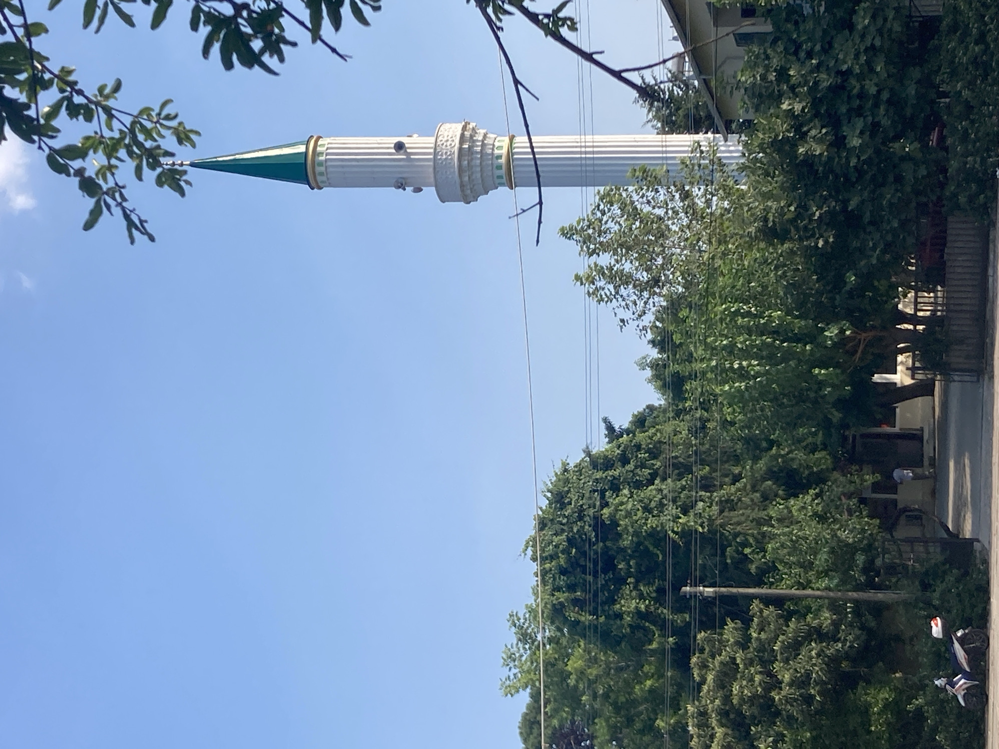 The image depicts a minaret of a mosque rising above lush greenery and a clear blue sky in the city of Kemer, Turkey. The minaret is tall and slender, with a white fluted shaft adorned with decorative bands of gold and green. It is topped with a conical green roof. In the foreground, dense trees and foliage create a natural barrier, with power lines crisscrossing overhead. A white scooter is parked on the left side of the frame. In the middle ground, a pathway leads to a building partially obscured by trees, where a person in a white shirt is visible. The overall scene suggests a tranquil setting in a residential or semi-urban area of Kemer.