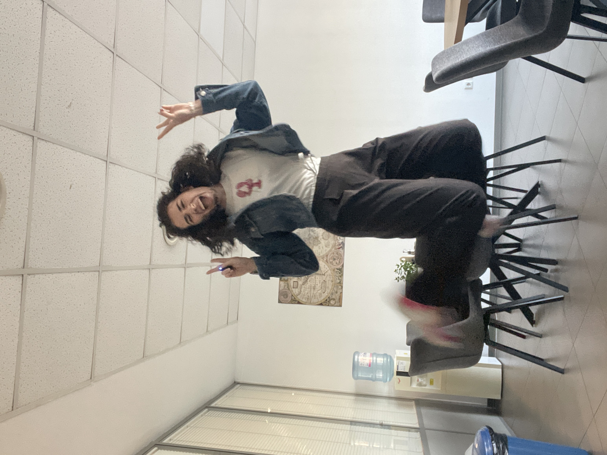 A dynamic, rotated photograph captures a young adult female mid-air, seemingly jumping or falling sideways, in an indoor office environment in Sofia, Bulgaria. The image is oriented 90 degrees clockwise, giving the impression that the subject is defying gravity.
The central figure has long, dark, curly hair and a wide, joyful smile, with her mouth open. She is dressed casually in a white t-shirt featuring a prominent red lobster graphic, dark-colored trousers, and a blue denim jacket. Her body is expressive, with bent arms and legs, and her right hand is raised with fingers spread. In her left hand, she holds a small, possibly glowing, blue object between her fingers. Motion blur around her feet suggests swift movement.
The background details an office setting: a white false ceiling with a visible grid pattern, plain white walls, and several dark grey office chairs with black metal frames. A water cooler with a large blue bottle stands against a wall, and a rectangular wall hanging, possibly an old map or decorative artwork, is affixed nearby. A blue trash can with a black liner is partially visible at the bottom of the frame. The presence of what appear to be glass partitions or windows at the bottom further defines the indoor space. The lighting is bright and evenly distributed, characteristic of an artificially lit interior.
The photograph conveys a strong sense of joy and playful energy, capturing a spontaneous and lighthearted moment within a professional setting.