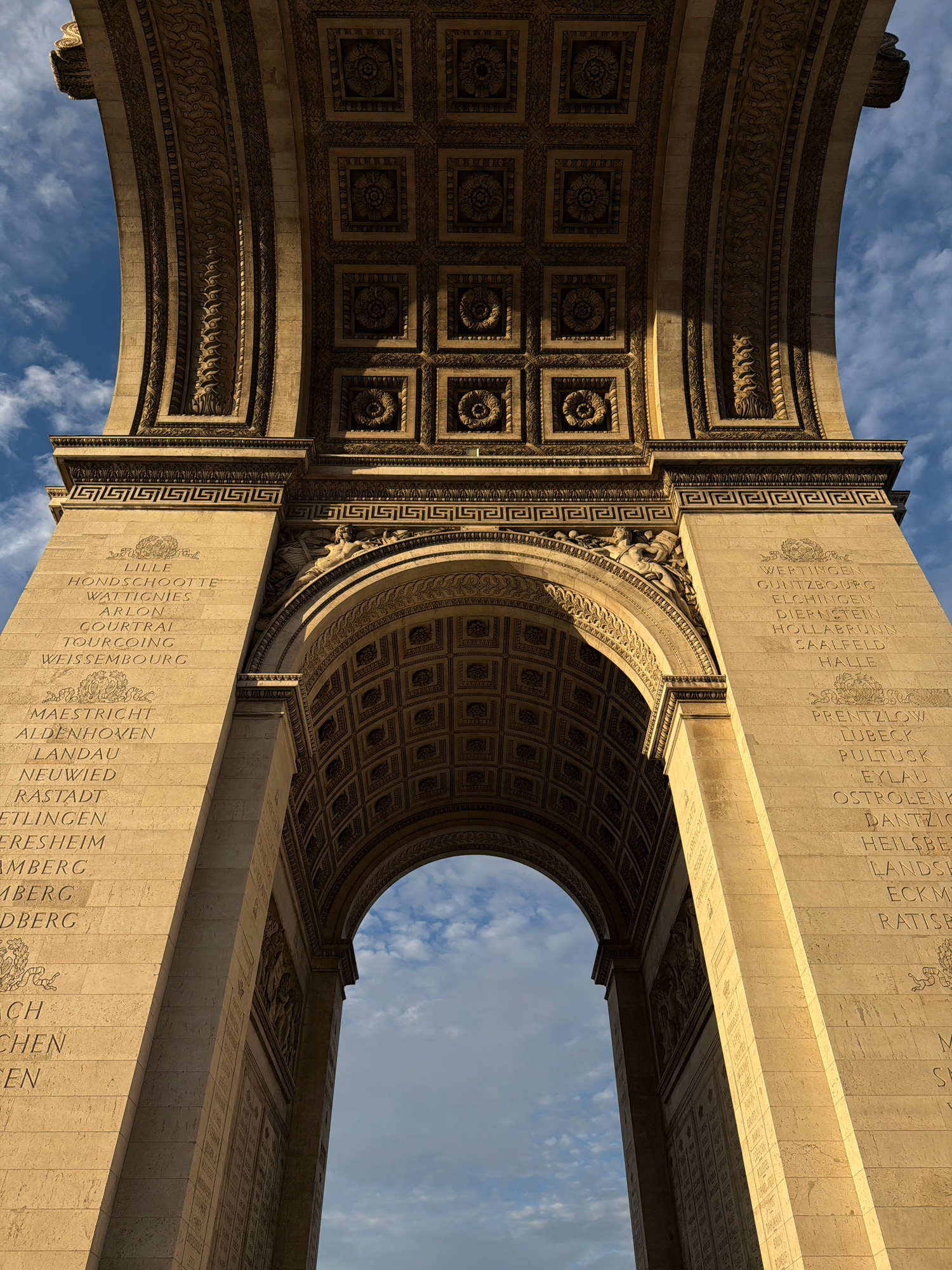 The image shows a low-angle, worm's-eye view of the Arc de Triomphe in Paris, France. The monumental arch is constructed of stone and features intricate carvings and inscriptions on its pillars and soffit. The sky, with scattered clouds, is visible through the archway and above the structure. The sunlight illuminates one side of the arch, casting shadows on the other, suggesting it is either morning or late afternoon. The names of battles and generals are inscribed vertically on the pillars of the arch, a common feature of this historical monument. No people are visible in the frame.