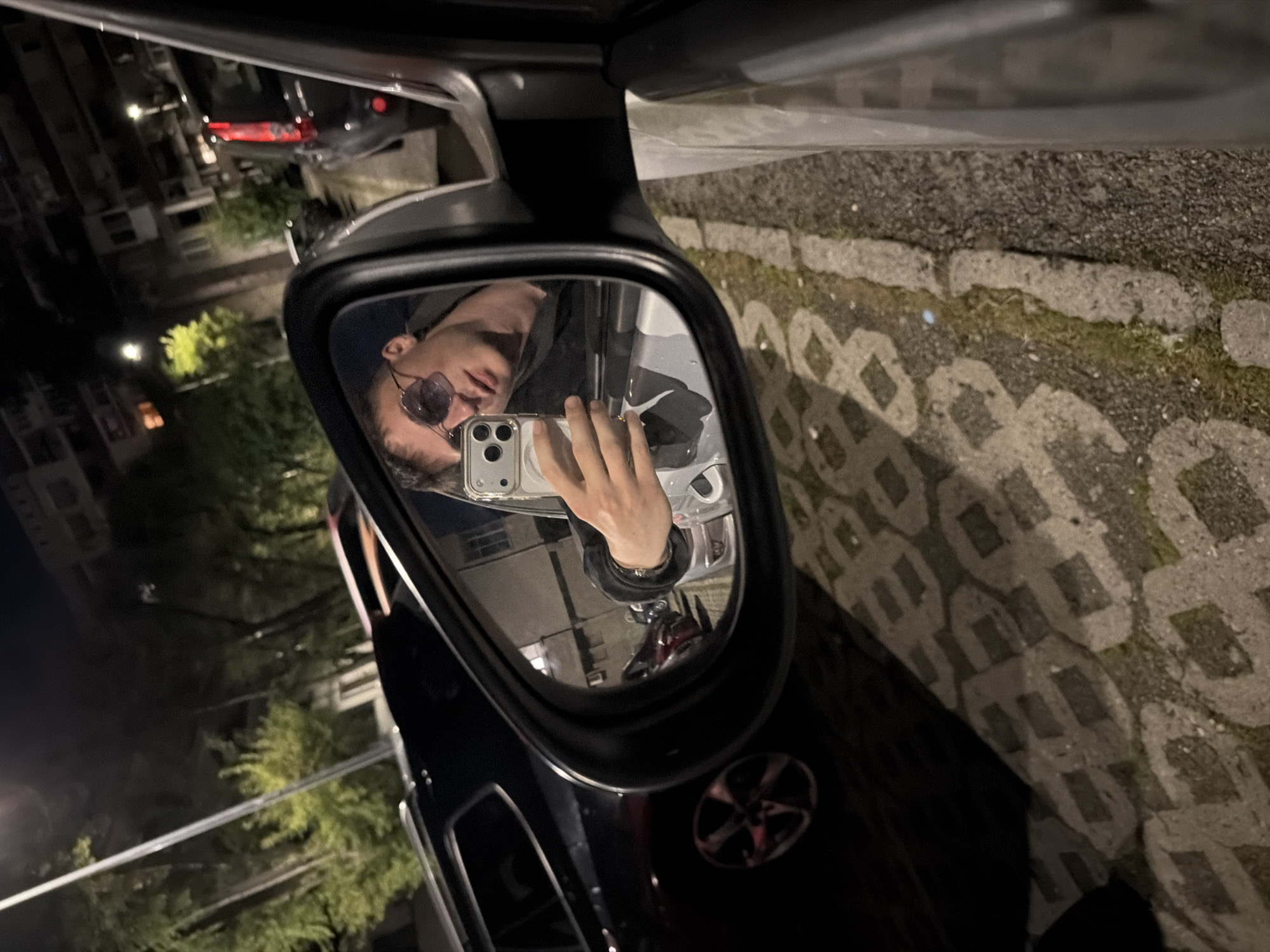 A man is reflected in a car's side mirror at night. He is wearing sunglasses and holding a smartphone. The street is paved with patterned concrete blocks. Buildings and street lights are visible in the background. The location is Burgas, Bulgaria.