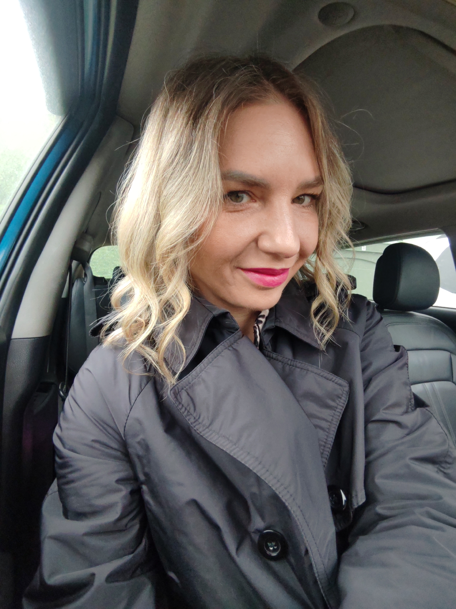 A woman is captured in a close-up selfie, seated inside a car. She has blonde, wavy hair, light skin, and striking green or hazel eyes. Her lips are adorned with bright pink lipstick, and she offers a pleasant, soft smile directly at the camera. She is wearing a dark grey or black trench coat, featuring a prominent collar and large black buttons on the front.
The setting is the interior of a car, indicated by the dark leather-like seats, the door frame, and the car's headliner, which appears dark grey and shows a circular fixture, possibly a light or part of a sunroof mechanism. Through the car windows, a blurry outdoor environment is visible, suggesting trees or greenery, along with a hint of a structure. The lighting appears soft and diffused, possibly indicating an overcast day. The overall impression is casual and relaxed. No visible text is present in the image.