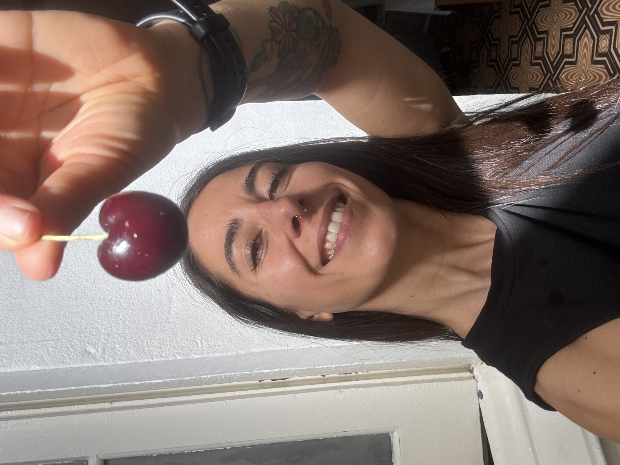 A woman is holding a single cherry above her head, with the cherry positioned directly over her forehead. She is smiling and looking directly at the camera. The woman has long, dark hair, a nose ring, and visible tattoos on her arm. She is wearing a black top. The background consists of a textured white wall and a patterned floor. Sunlight is casting shadows across the scene, suggesting an outdoor or well-lit indoor environment. The image was captured in Barcelona, Spain.