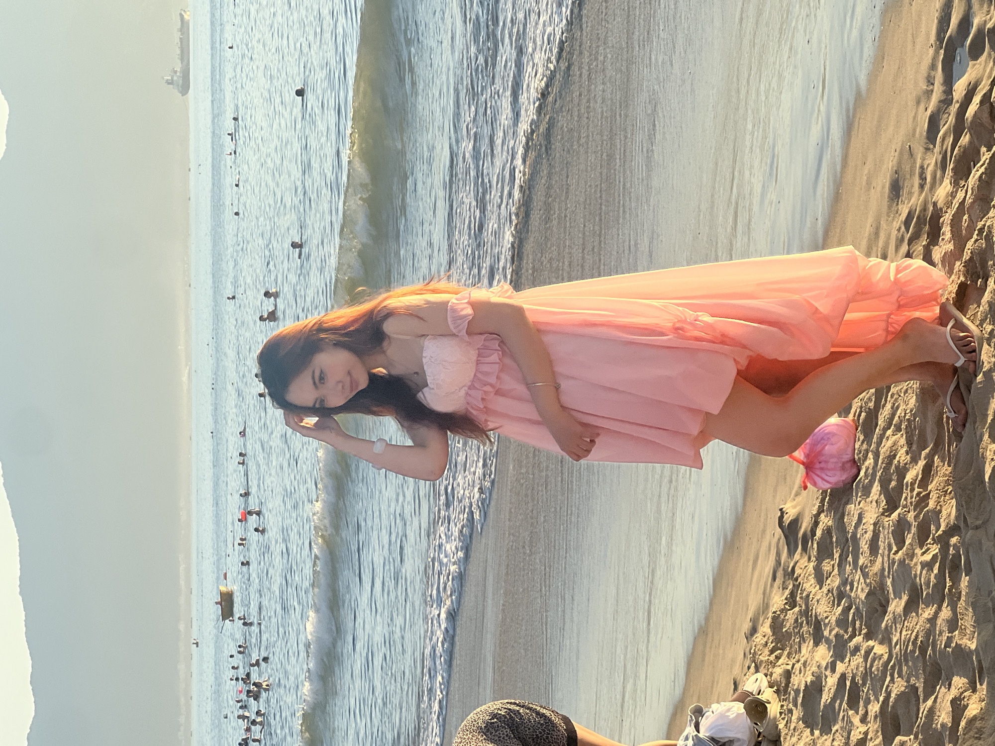 In Đà Nẵng, Vietnam, a young woman stands on a sandy beach with the ocean behind her. The water is calm, with small waves reaching the shore. Numerous people are visible in the water, some swimming and others wading. A small boat with a person in it is further out in the sea, and a large cargo ship can be seen on the horizon. The sky is hazy, with mountains in the distance. The woman is wearing a flowing pink dress and is posing with her hand on her head. Her feet are bare and she is wearing flip-flops. A pink bag is on the sand near her. The lighting suggests it is late afternoon or early evening.