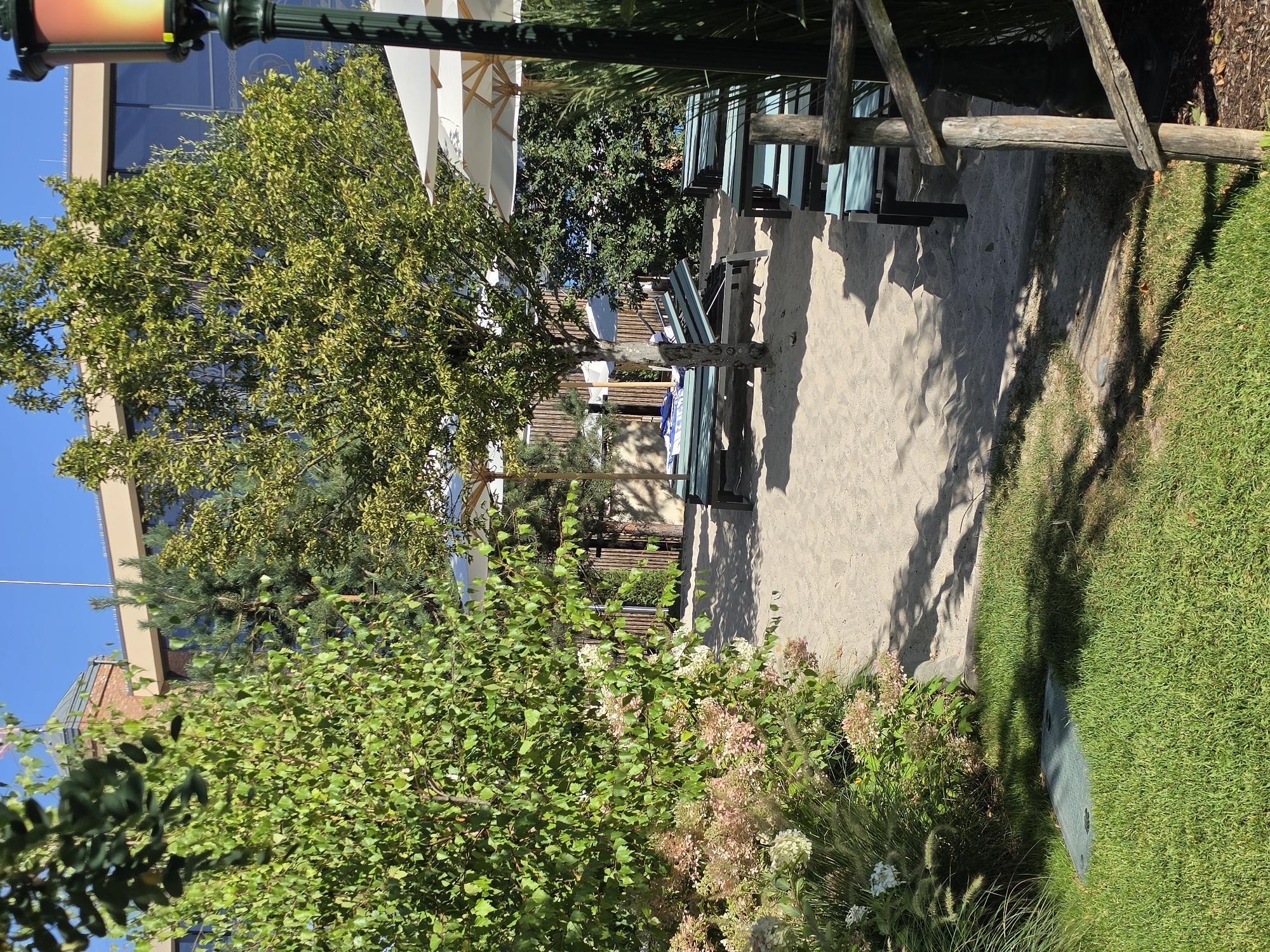 The image depicts an outdoor seating area with sand, lounge chairs, and umbrellas, suggesting a beach or a poolside relaxation zone. A building with a modern facade and large windows is visible in the background, partially obscured by trees. The scene appears to be a well-maintained public space, possibly part of a resort or a recreational facility. The lighting and shadows suggest it is daytime, with the sun casting distinct patterns on the sand and grass. The presence of lush greenery, including trees and flowering bushes, adds to the serene atmosphere.
This scene is located at the Europa-Park Hotel "Bell Rock" in Rust, Germany. Specifically, it is the beach area adjacent to the hotel, offering guests a resort-like experience with sand, sun loungers, and a view of the hotel's distinctive architecture. The hotel itself is themed around a lighthouse and maritime elements, which is subtly reflected in the design of the outdoor space.