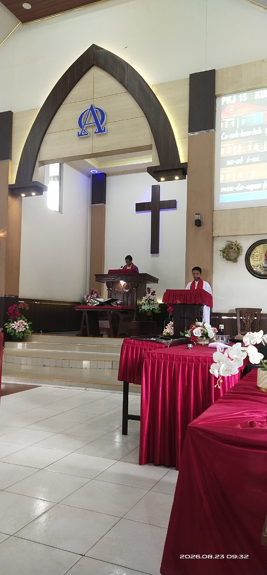 The image depicts the interior of a Christian church, likely in Jaar, Indonesia, given the timestamp. Two young men in white shirts and red stoles are positioned at separate podiums, suggesting they are involved in a religious service. One man stands at a larger, ornate wooden pulpit on a raised platform, facing left, while the other stands at a smaller, draped podium in the foreground, facing forward. A large wooden cross is mounted on the wall behind the first man. The room features a light-colored tiled floor, white walls, and a wooden ceiling with recessed lighting. A screen displaying text, possibly song lyrics or scripture, is visible on the right side of the image. In the foreground, two tables are covered with vibrant red cloths, adorned with floral arrangements. The overall atmosphere appears to be one of a solemn religious gathering.