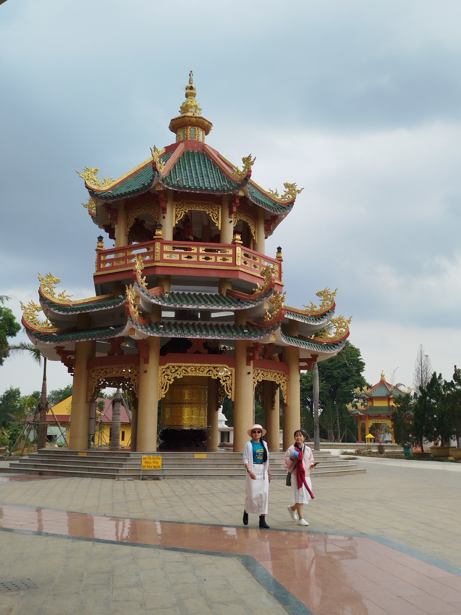 Two women are walking in front of a large, ornate, pagoda-like structure in Phú An, Vietnam. The structure has multiple tiers with green roofs and gold detailing. A large golden bell hangs in the center. One woman wears a white skirt and jacket, a blue t-shirt, a hat, and boots. The other woman wears a white skirt and sneakers with a pink jacket draped over her shoulders. The plaza is paved with patterned tiles. Another, smaller pagoda is visible in the background. The sky is overcast.