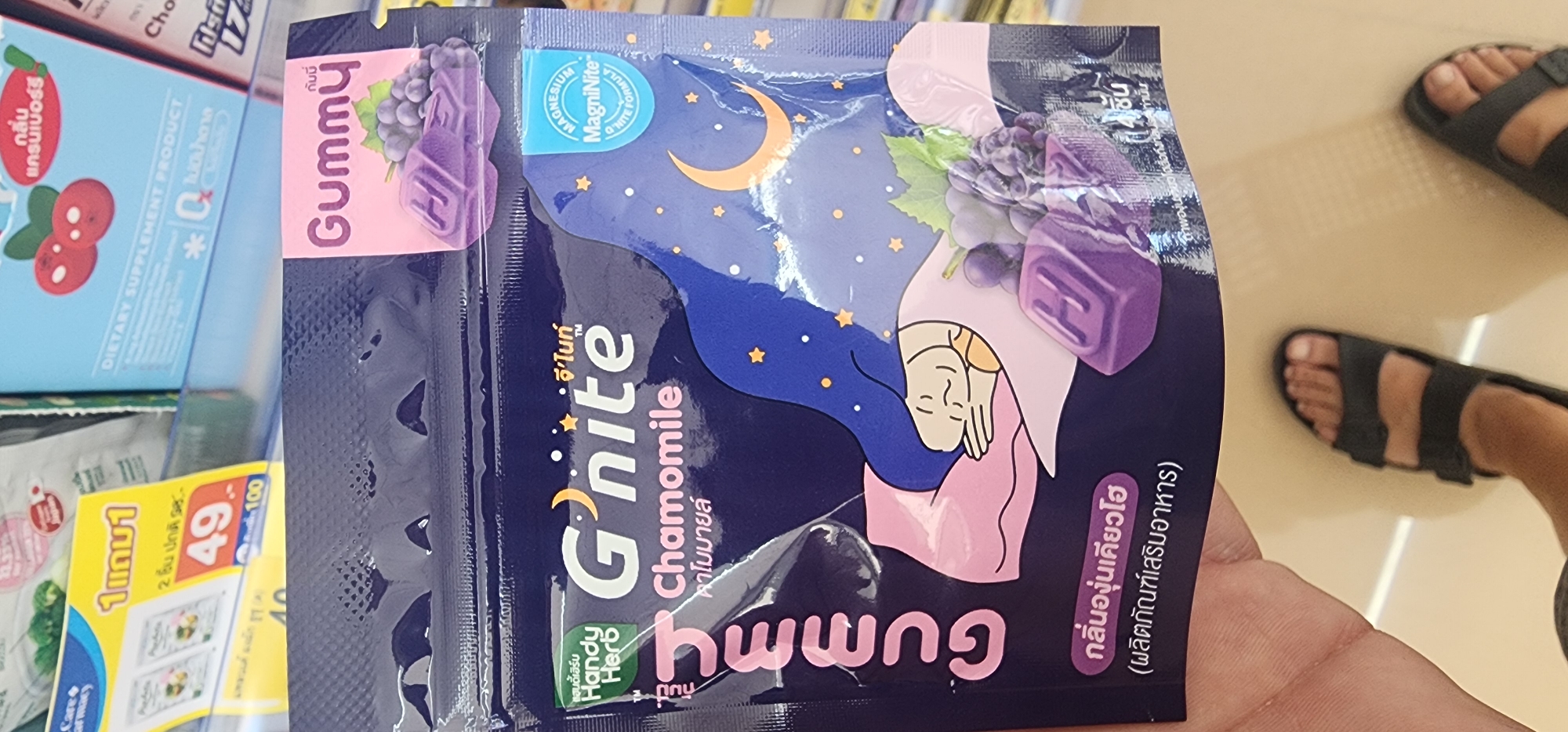 This is a close-up, eye-level, first-person perspective photo of a person's hand holding a pouch of "G'nite" brand gummy supplements. The pouch is dark purple and features an illustration of a person sleeping peacefully, surrounded by stars and a crescent moon, and a cluster of purple grapes with two purple, cube-shaped gummies. The text on the pouch is in Thai and English, with "G'nite" and "Chamomile" being prominent. A small blue tab on the upper right of the pouch contains text in English and the brand name "Magnite". The background shows blurred shelves with other products, consistent with a retail environment like a convenience store or pharmacy in Ko Samui District, Thailand. The person is wearing black sandals, with their feet visible at the bottom of the frame. The lighting is bright and even.