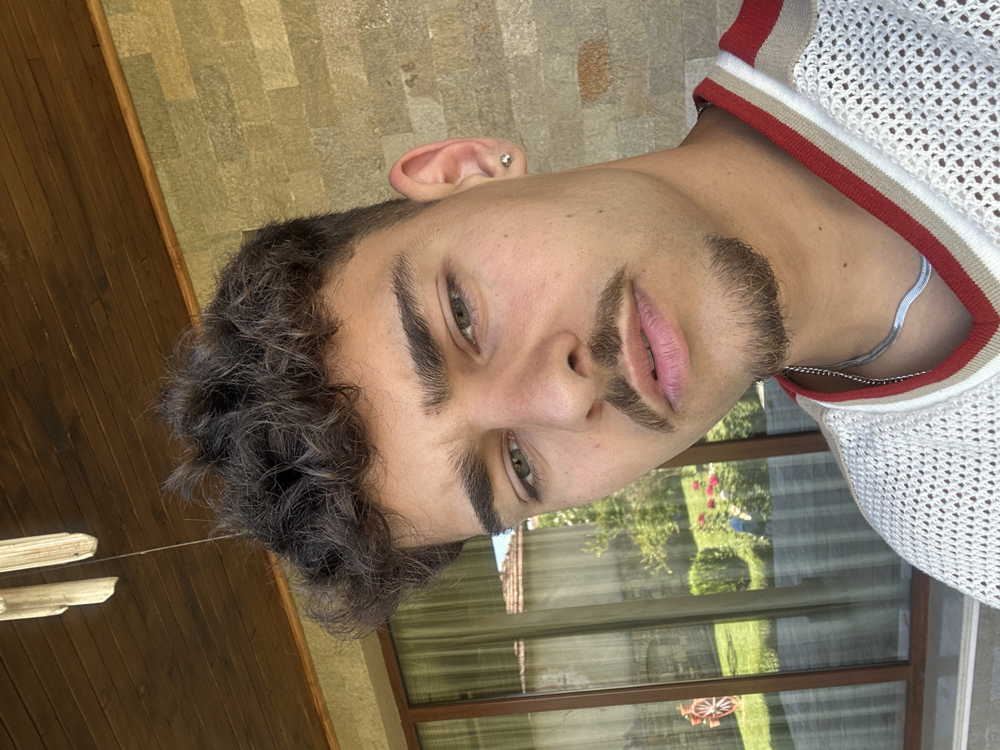 A young man with olive skin, dark curly hair, and green eyes is the focus of a vertically-oriented medium shot. He has a small earring in his left ear and a mustache and goatee. He is wearing a white mesh shirt with a red and beige stripe at the collar, and a silver chain is visible around his neck.
The background appears to be a building's exterior, with a wooden paneled wall on the left and a window reflecting a green lawn and what appears to be a child's toy wagon. The photo was likely taken in Dermantsi, Bulgaria.