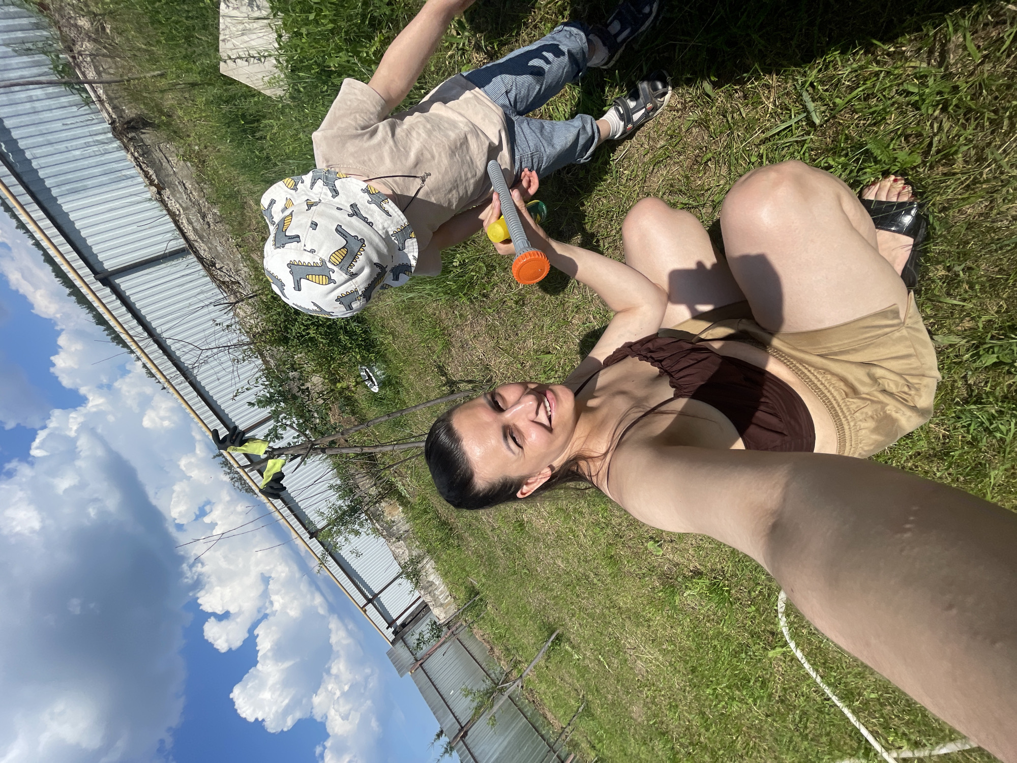 In Olkhovka, Russia, a woman with dark hair pulled back is sitting on the grass, taking a selfie outdoors. She is wearing a brown crop top and tan shorts. Beside her, a young child wearing a white hat with dinosaur prints, a light brown t-shirt, and blue striped pants is standing. The child is holding a toy that resembles a water gun. The background features a grassy yard with young trees, a corrugated metal fence, and a bright blue sky with fluffy white clouds. The overall scene suggests a casual, warm-weather activity in a rural or suburban setting.